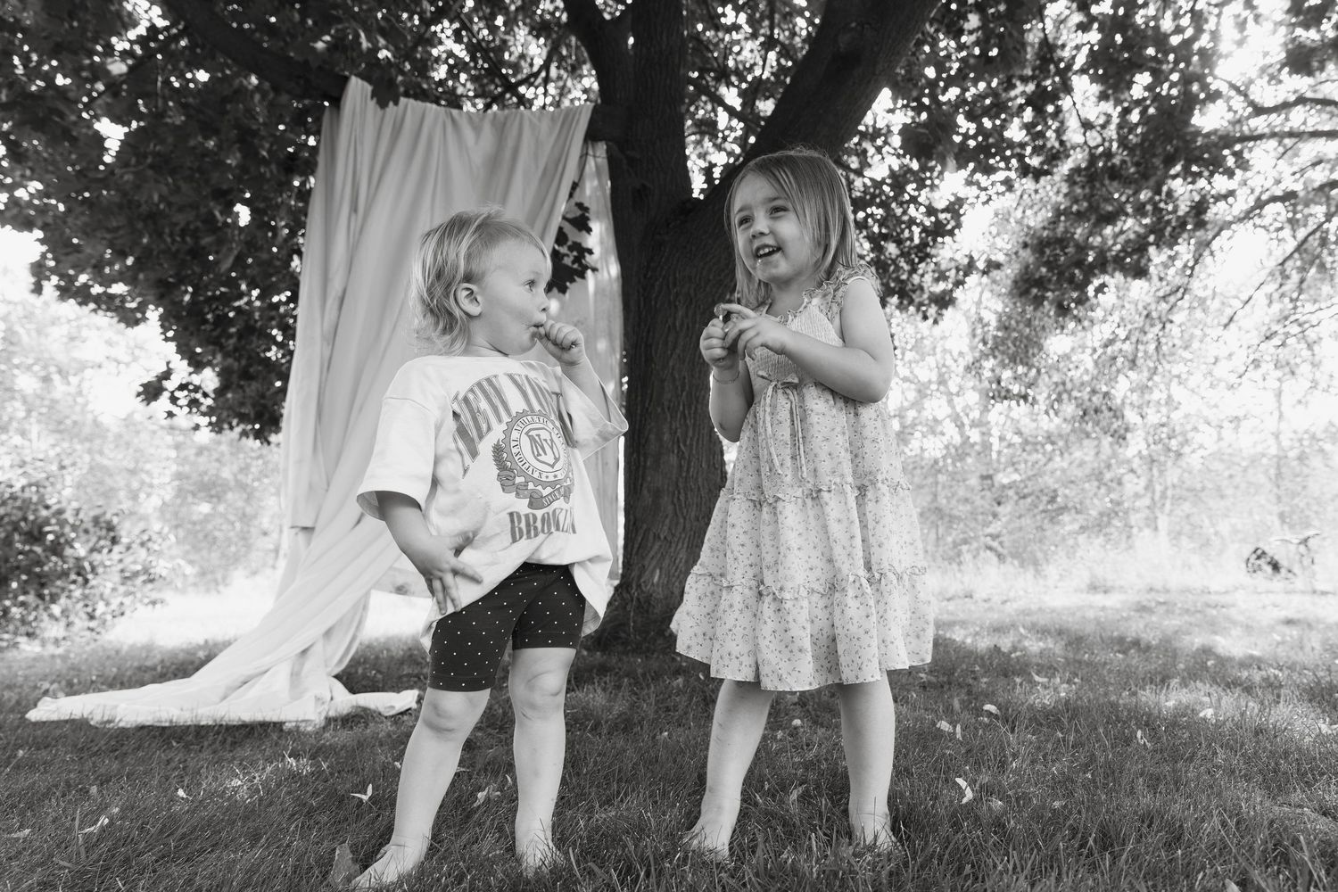 Children in white dresses playing under a large tree in a black and white photograph.