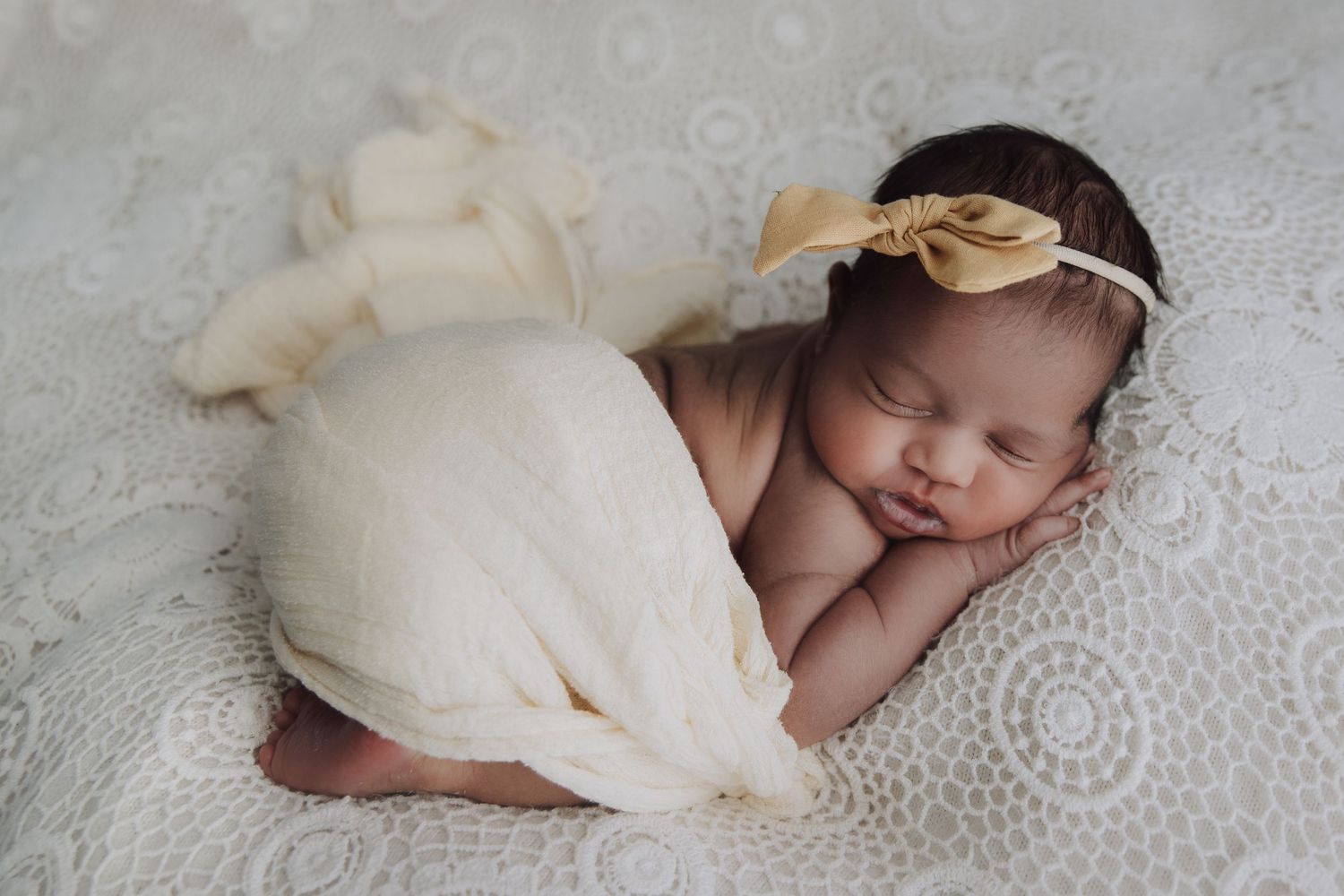 Sleeping newborn in cream wrap with gold leaf headband rests on textured white blanket.