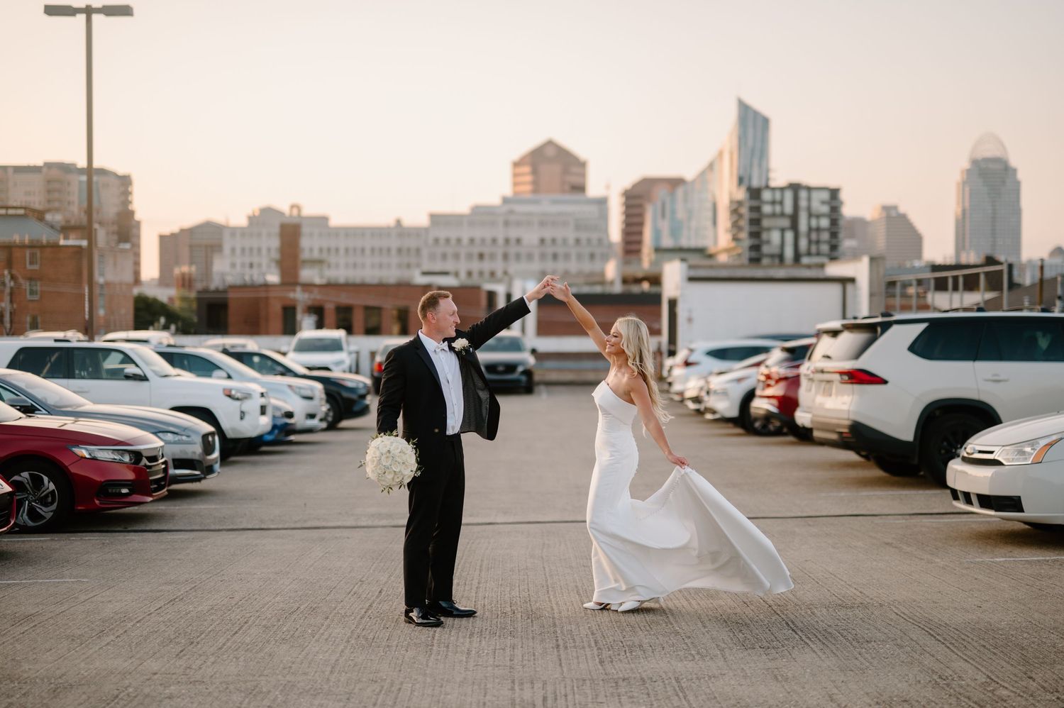 A wedding couple walks through a parking lot with city buildings visible in the background during sunset.