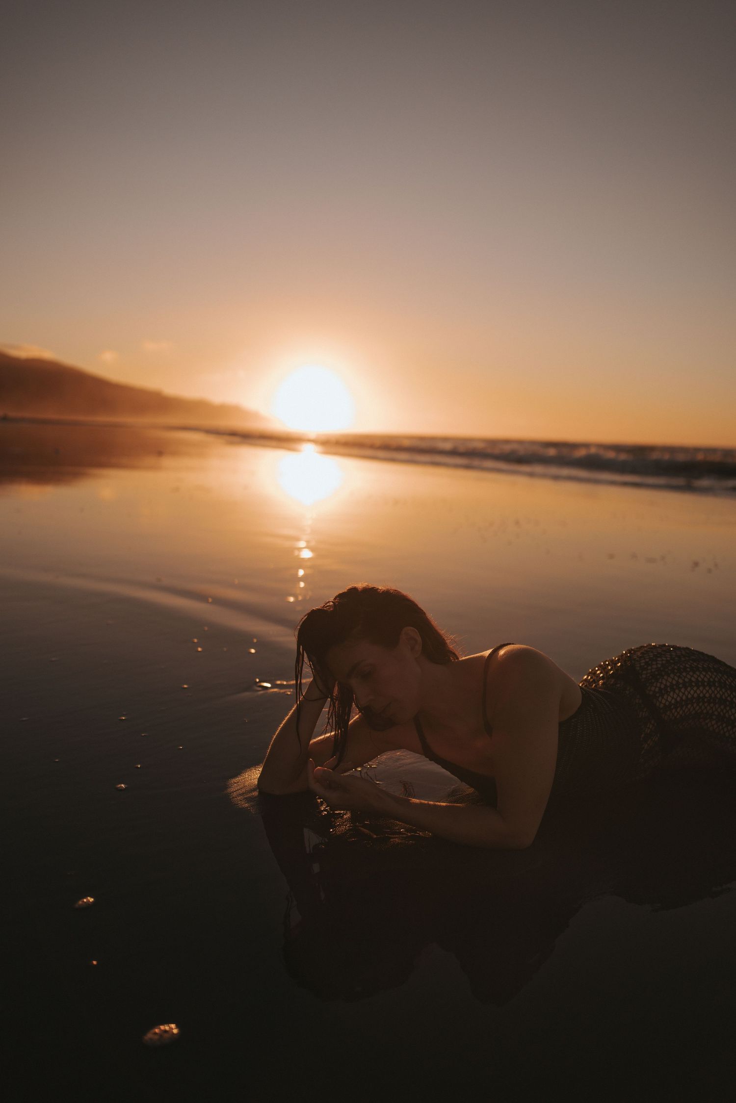 A woman lies on a wet beach at golden sunset, waves reflecting warm orange light across the shoreline.