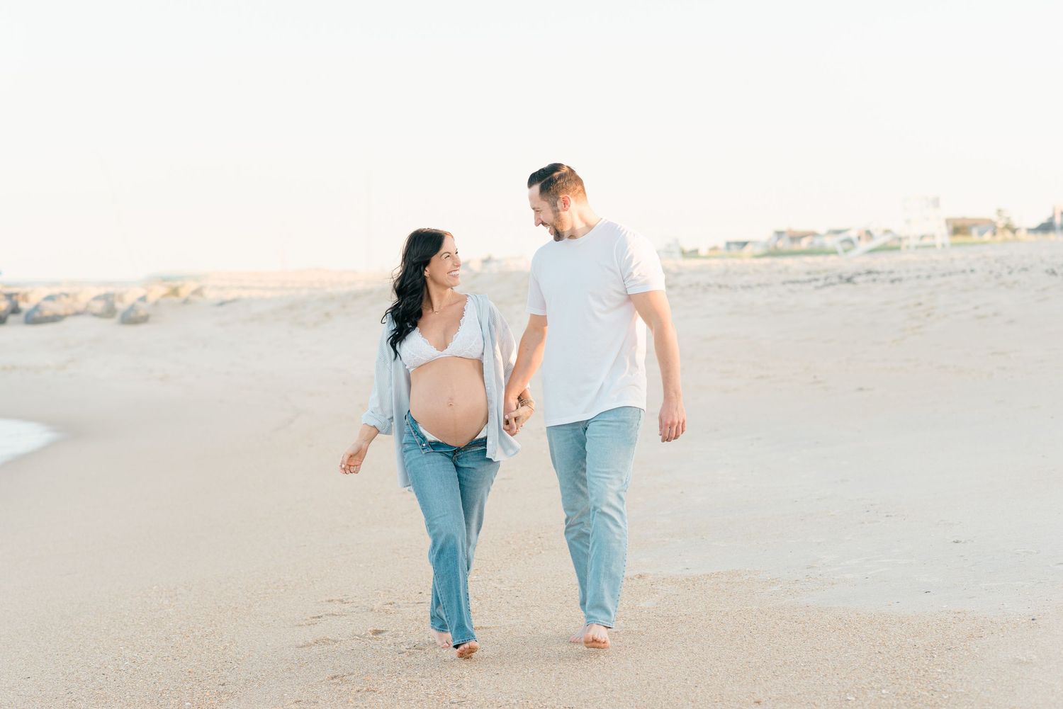 Candid maternity session of expecting mother walking down the jersey shore beach with her husband holding hands laughing.