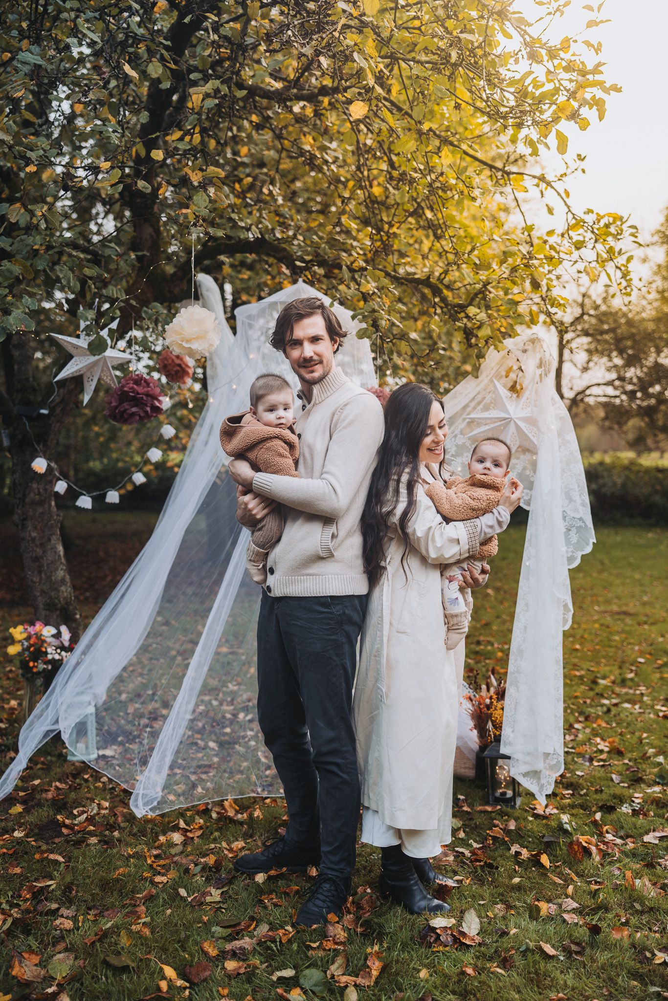 Young family with twin babies posing outdoors in autumn garden with drapings and fairy lights decorations and golden foliage.