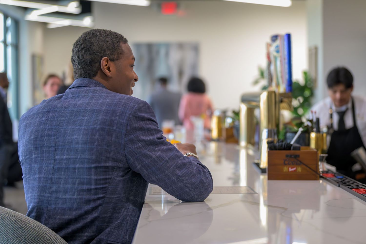 Man in blue plaid suit sits at a white counter bar while a chef works in the background.
