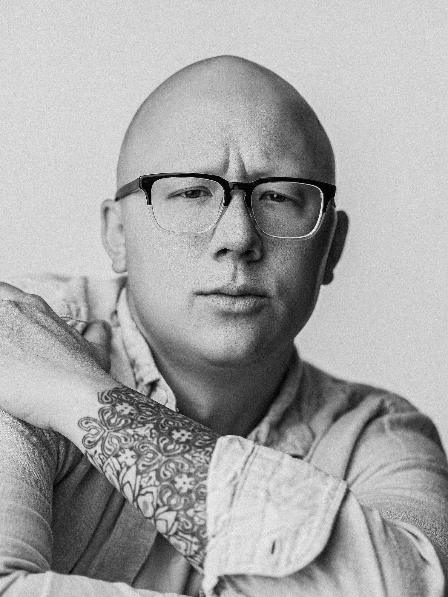 Bald man with glasses and floral tattoo sleeve poses for a black and white portrait photo.