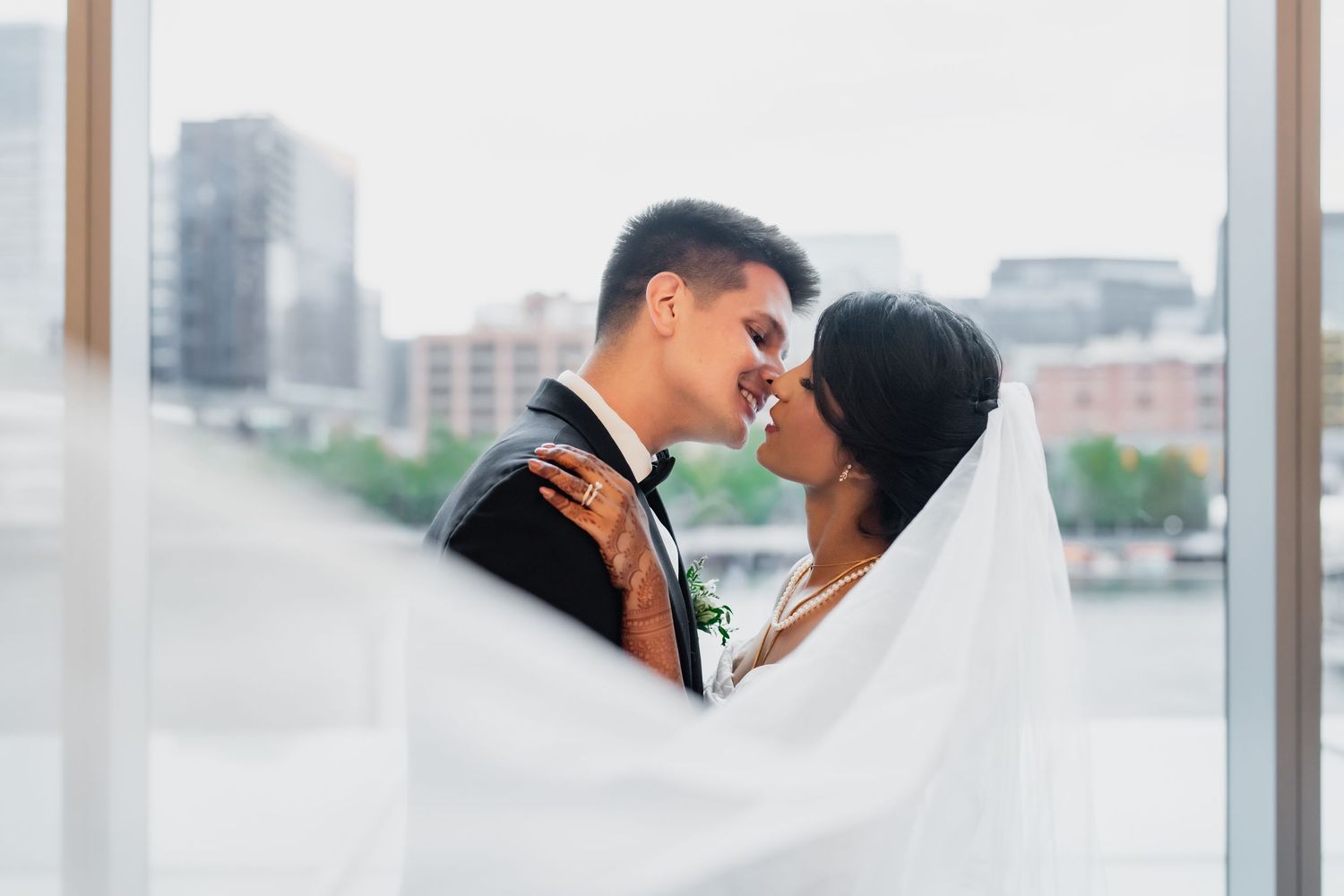 Bride and groom share romantic moment under veil with city skyline backdrop on wedding day.