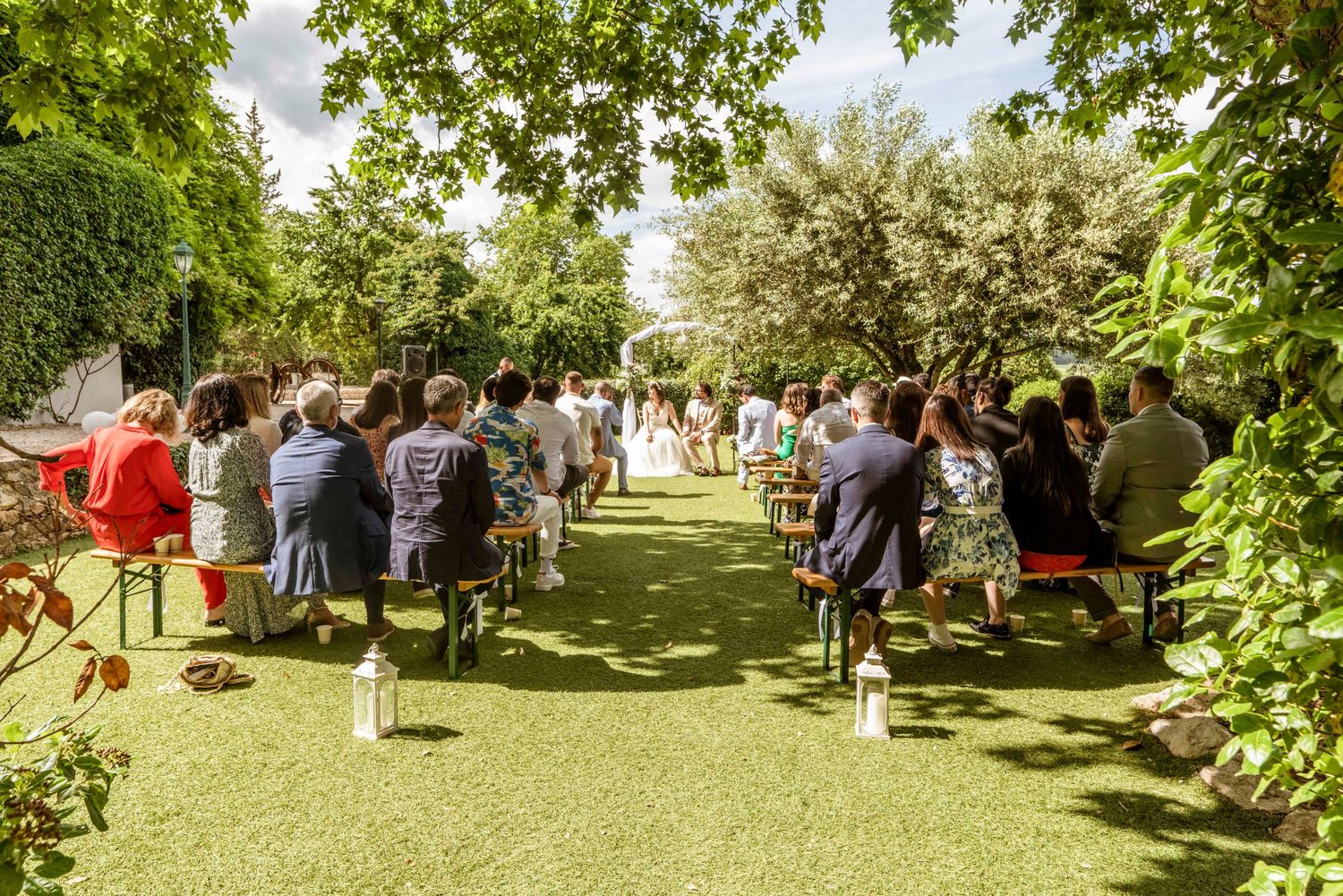 A panoramic view of a sunny outdoor wedding ceremony with guests seated in white chairs under lush green trees.