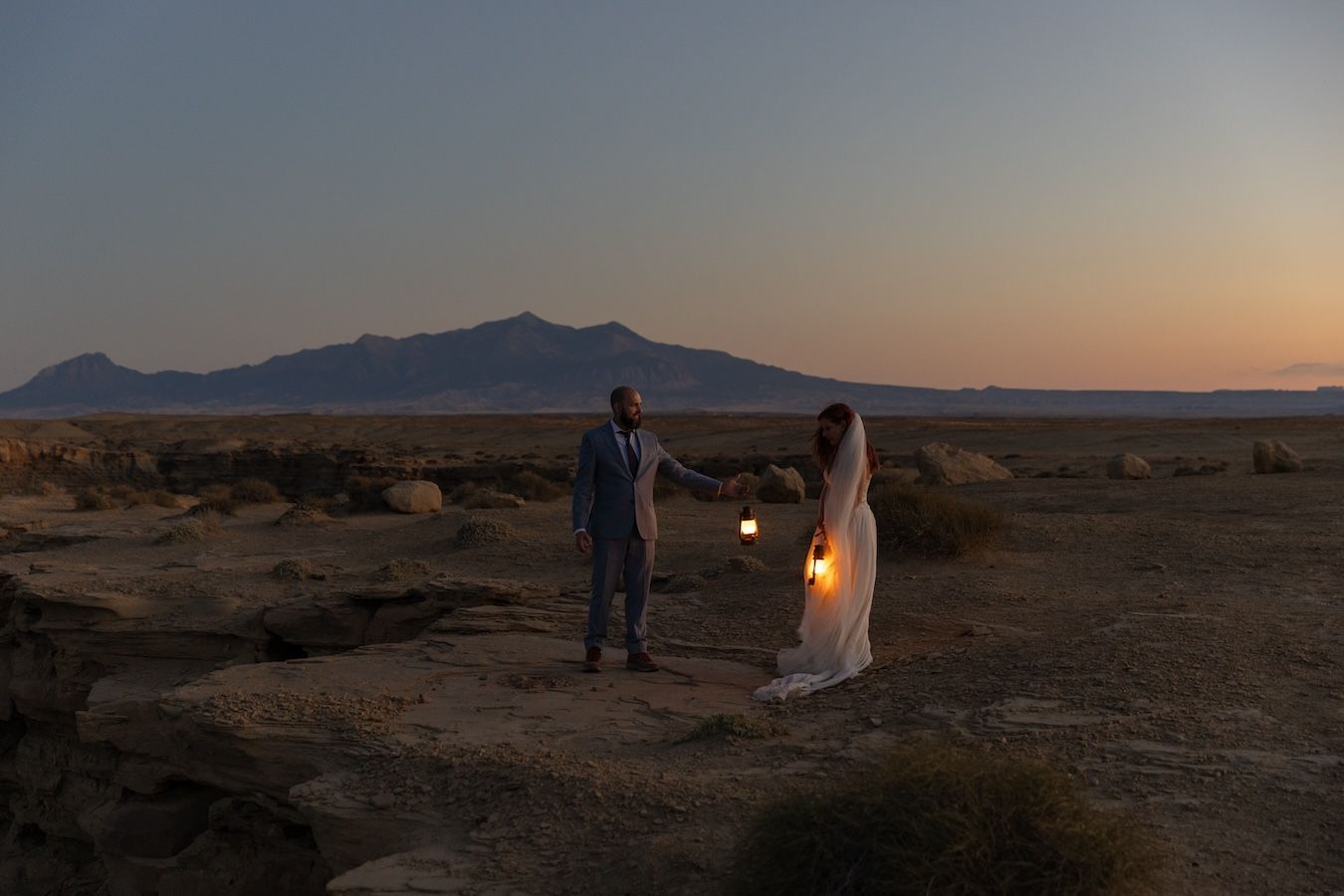 Two figures stand in a desert landscape at dusk with mountains in the background and a glowing lantern between them.