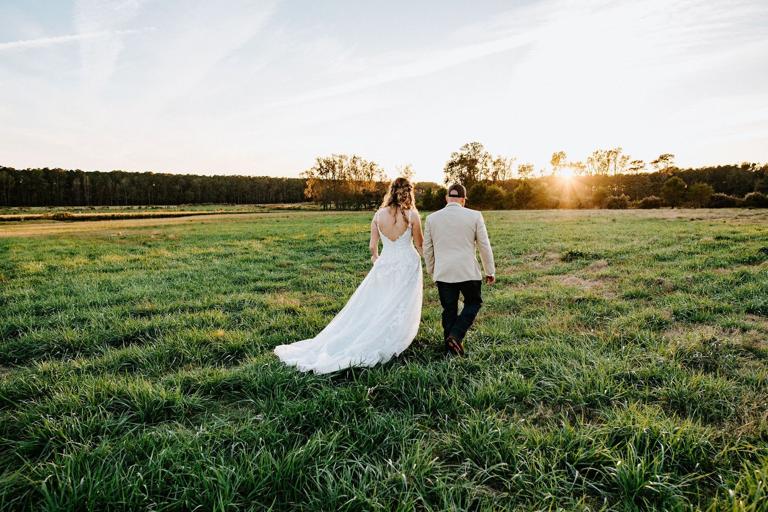 Couple walks hand in hand through a golden meadow at sunset as her white wedding dress trails behind them.