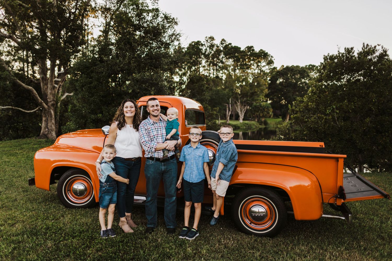 Family of six posing in front of a classic orange vintage pickup truck on a lush green lawn at sunset.