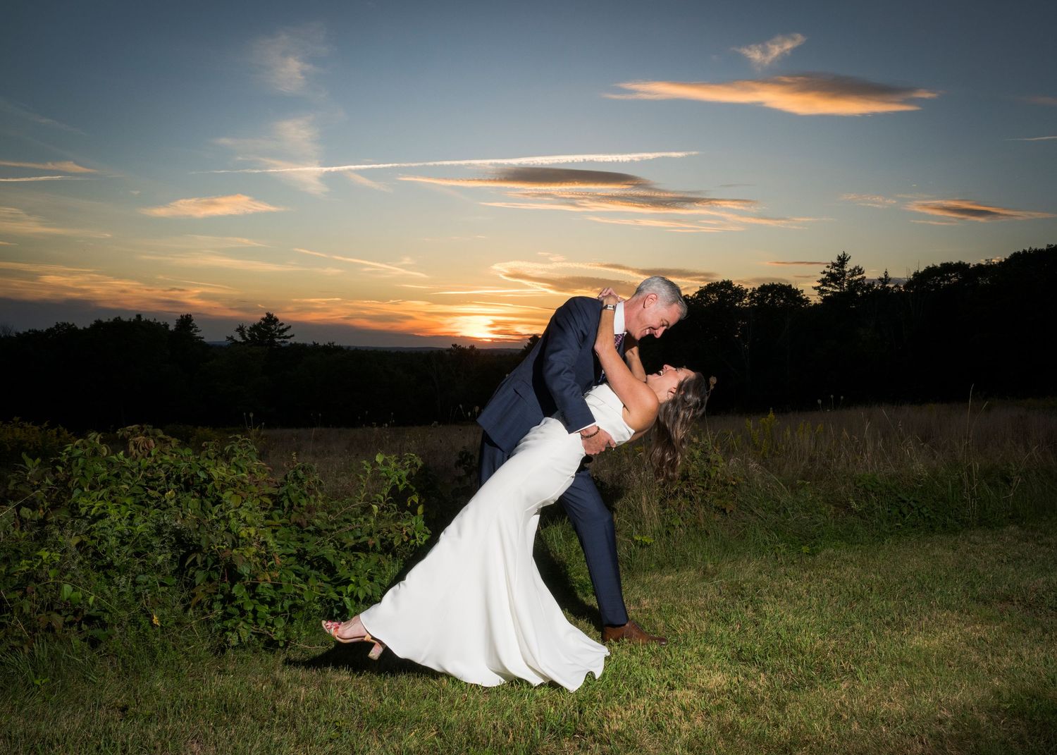 Bride and groom share a romantic dip kiss at sunset in a lush green field with a vibrant orange sky.