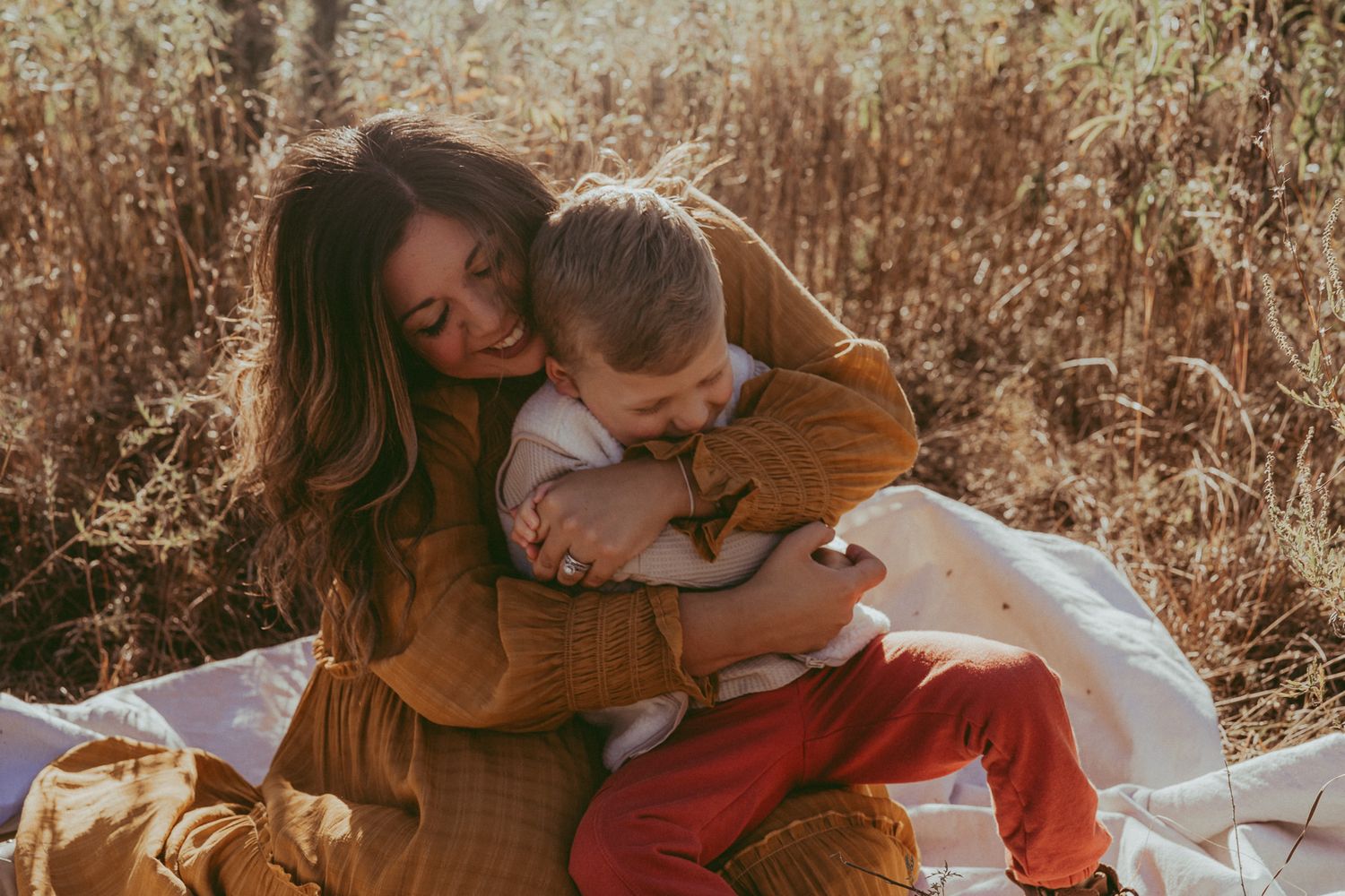 Family of four sitting on blanket in golden field, parents holding toddlers, warm autumn outdoor portrait.