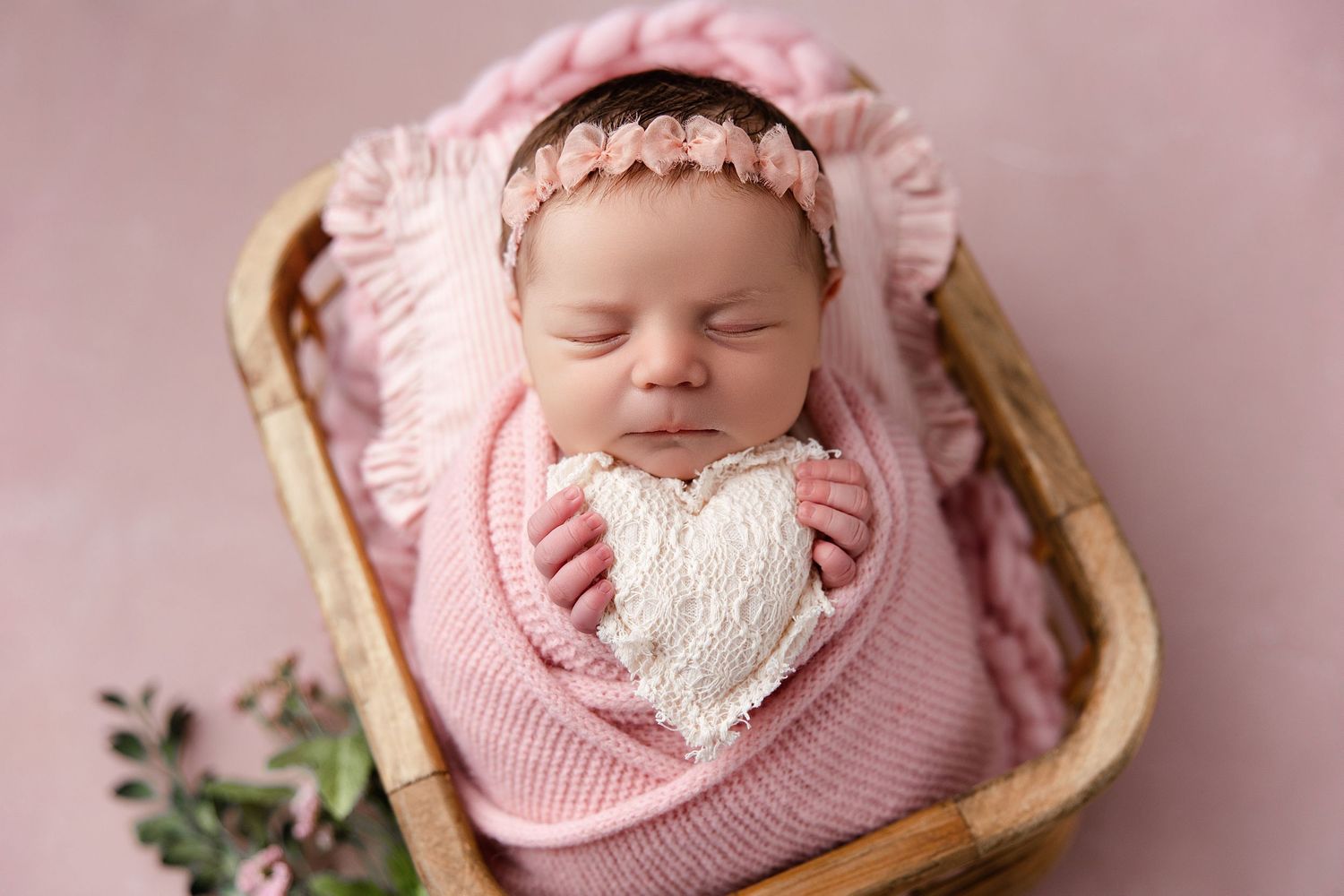 A sleeping baby in pink knit clothing lies in a wooden basket with floral decorations.