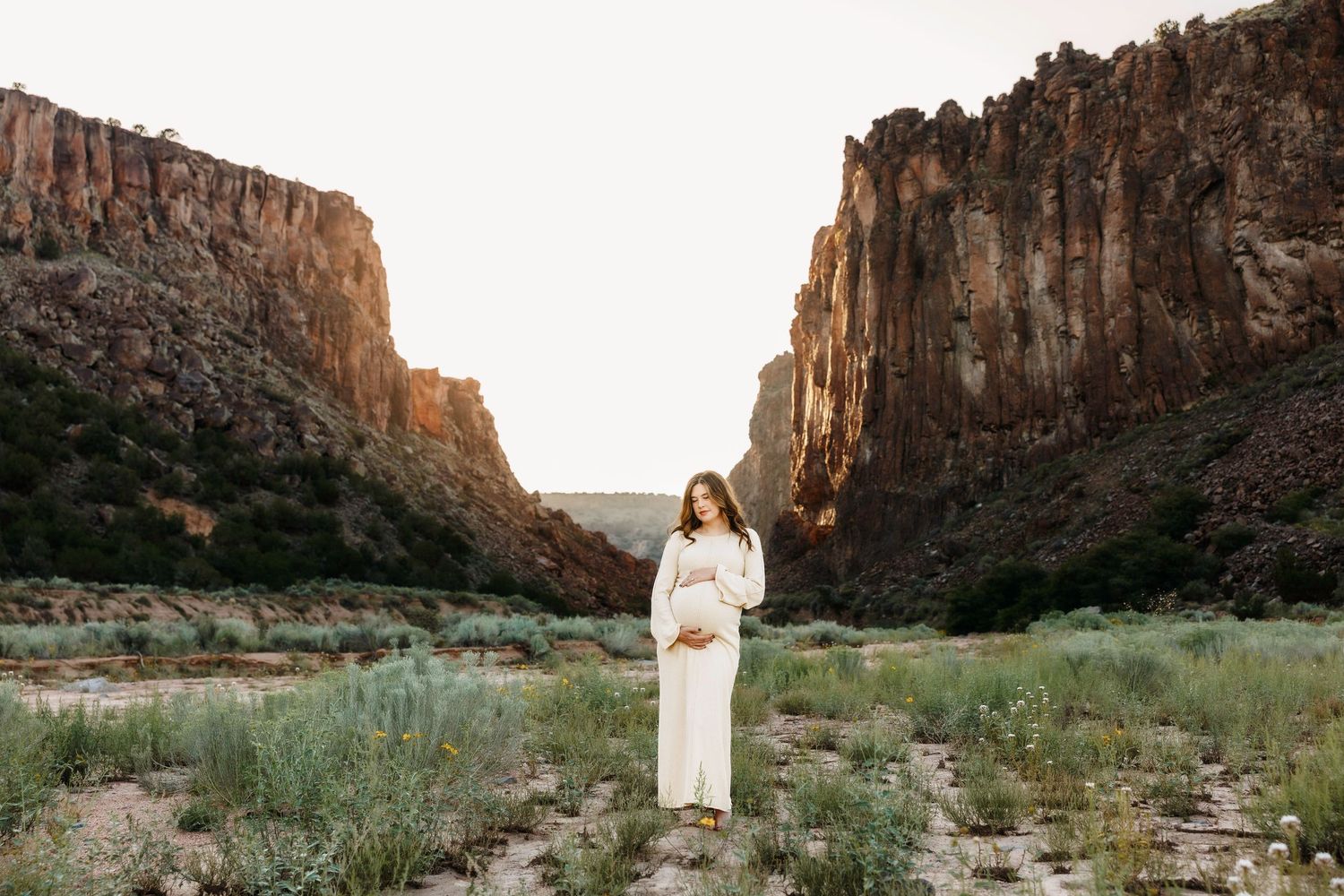 Pregnant woman in white dress standing in a desert canyon with rocky cliffs at golden hour.