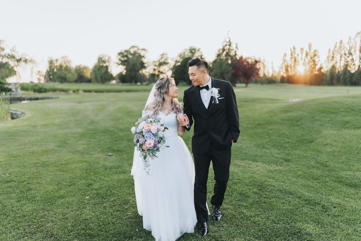 Newlywed couple walks together on lush green golf course during sunset holding beautiful purple floral bouquet.