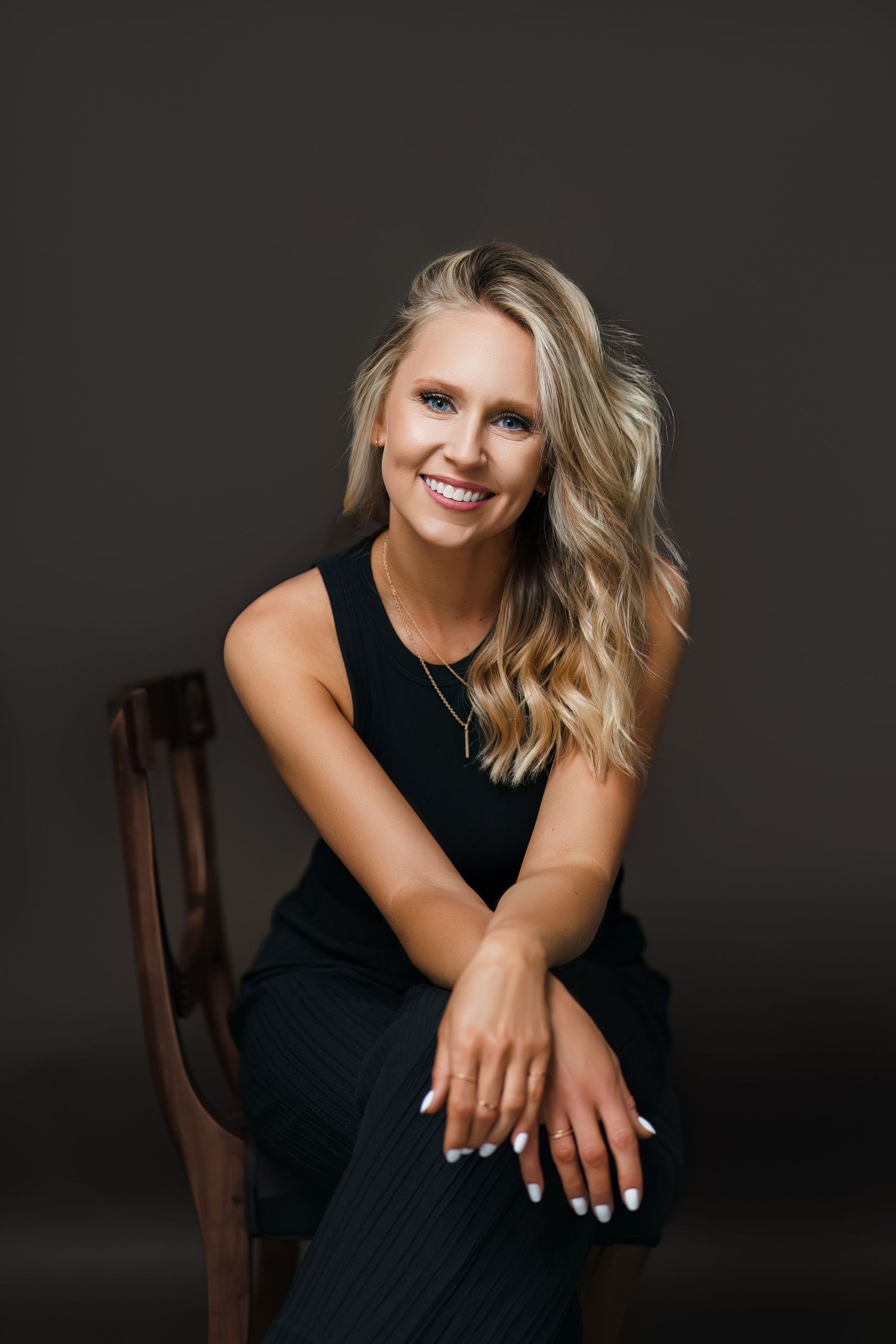 Portrait of someone wearing a black sleeveless dress seated on a wooden chair against dark background with blonde wavy hair.