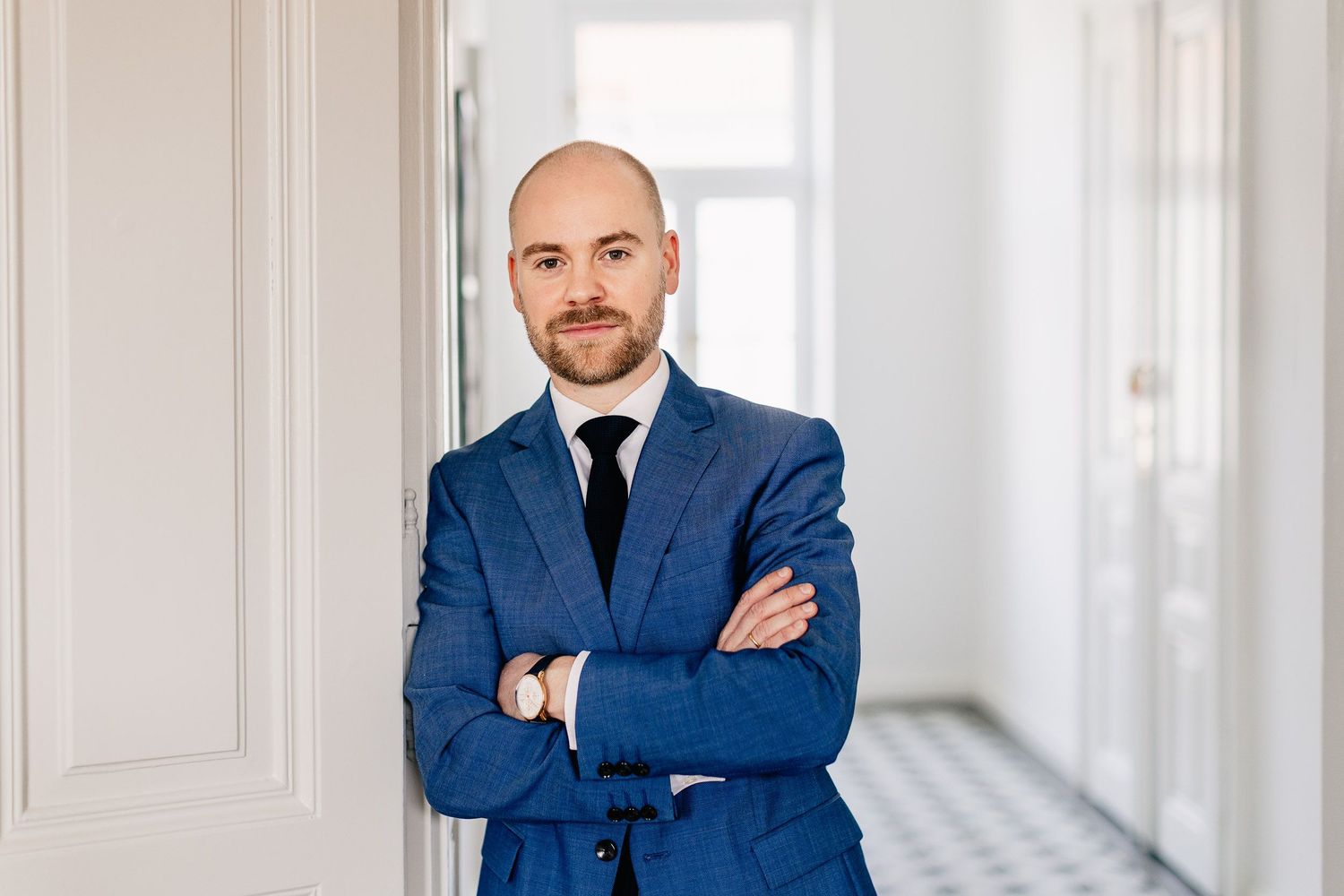Professional in a navy blue suit stands with arms crossed in a bright white hallway with architectural columns.