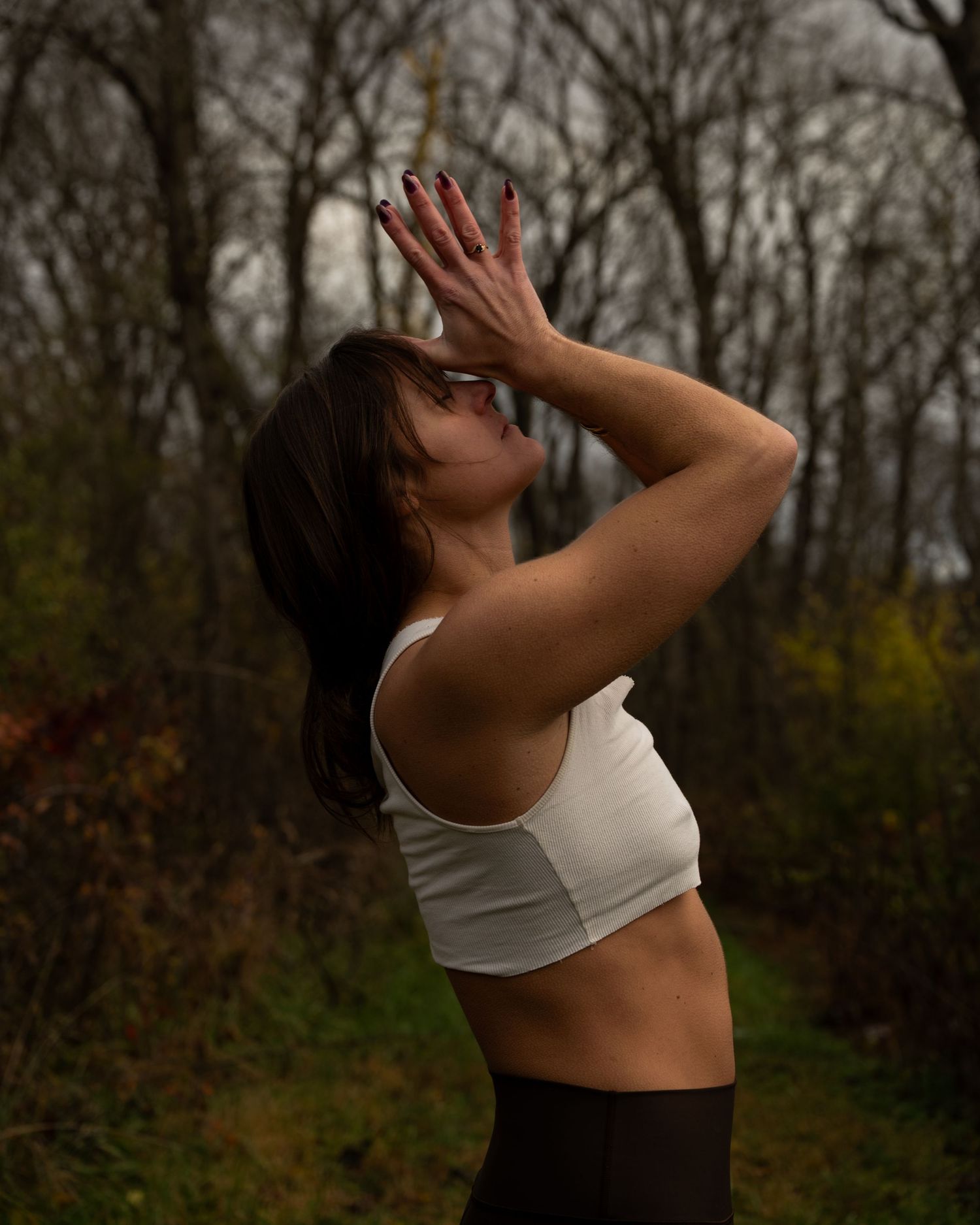 Athletic woman in white crop top stretches outdoors with raised arms against moody forest backdrop.