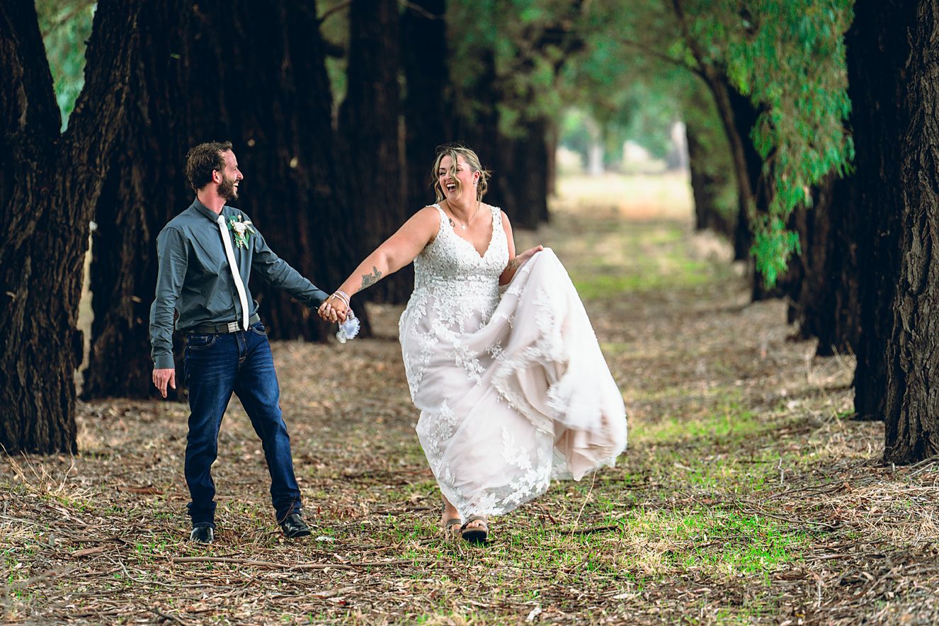 A couple in wedding attire walks through a wooded area during an outdoor photo session.