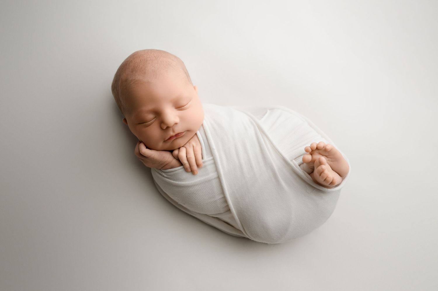 Sleeping newborn baby wrapped in white swaddle blanket photographed on light gray background.