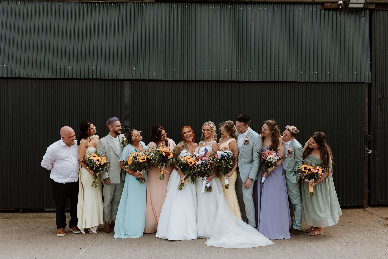 A small group poses for a wedding photo outdoors with a white dog on a stone wall backdrop.