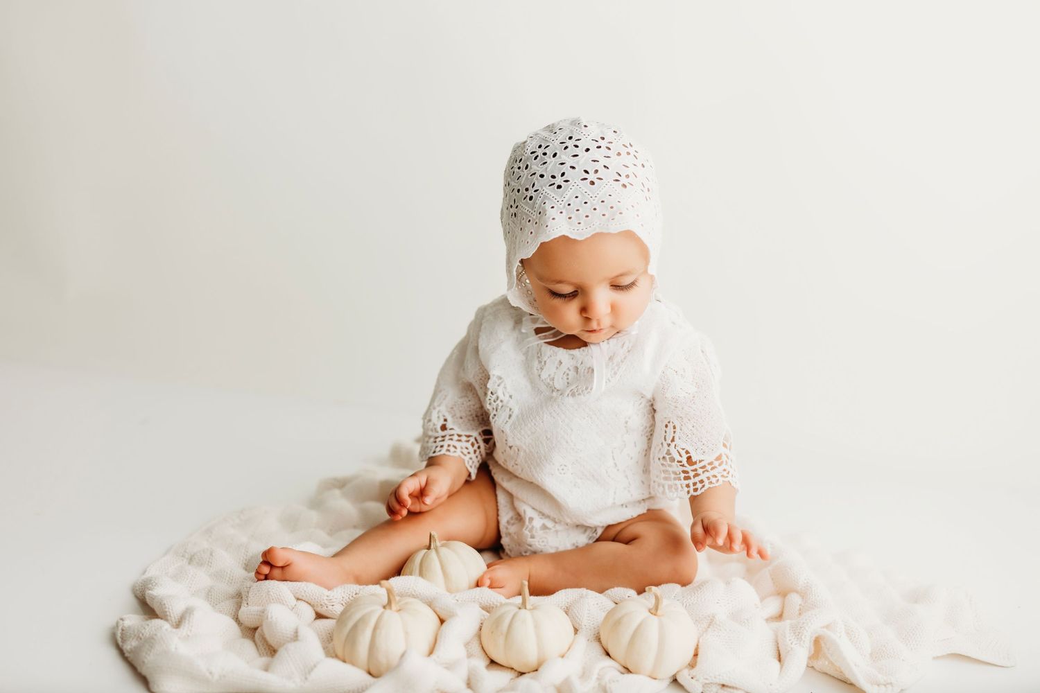 Adorable baby in white lace bonnet sits among soft white blankets and pillows in dreamy nursery setting.