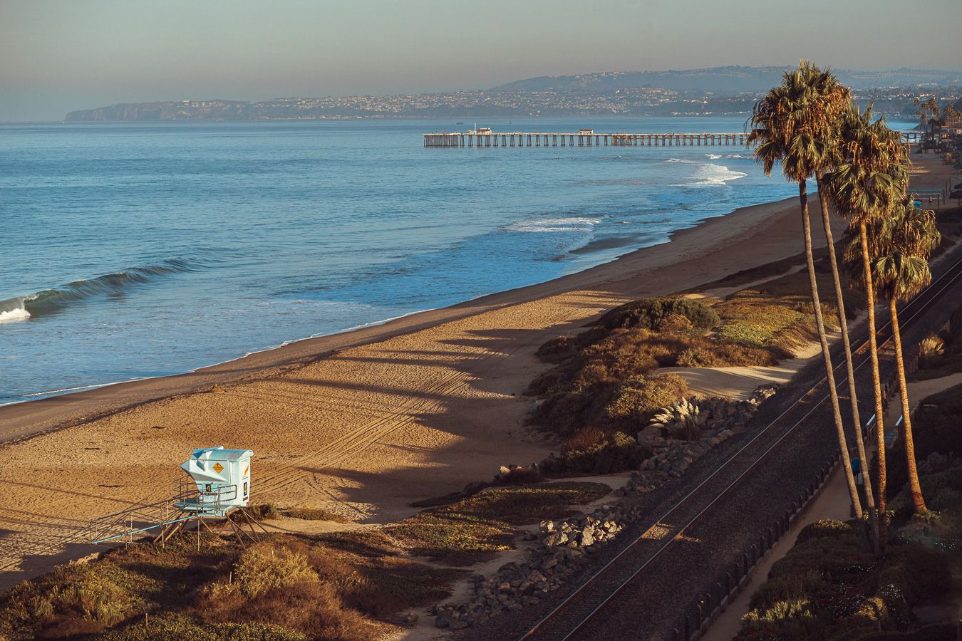 Scenic coastal beach view with tall palm trees, lifeguard tower, and rolling waves along the California shoreline at sunset.