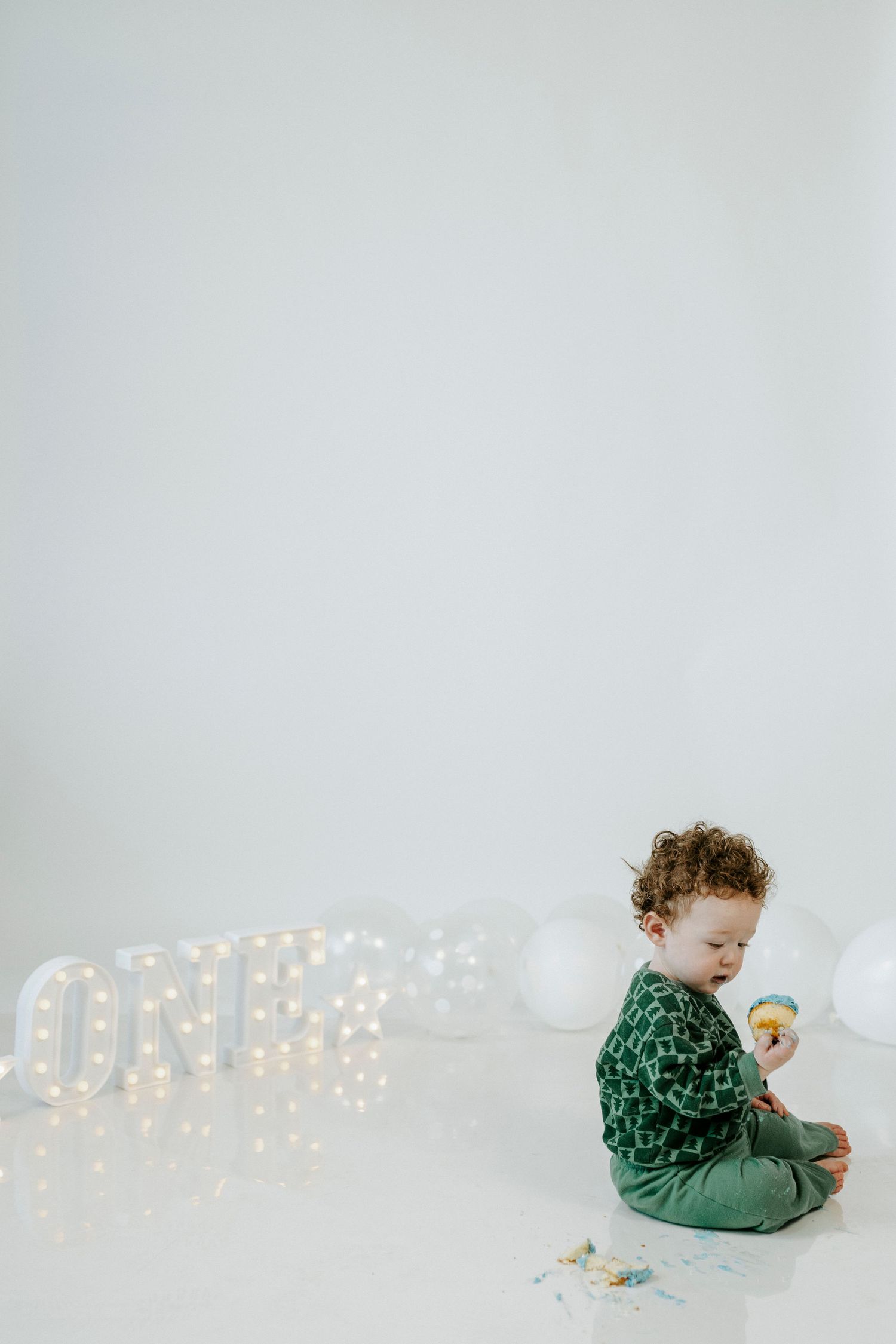 Baby in green outfit sits on white backdrop with illuminated letter lights and scattered confetti decorations.