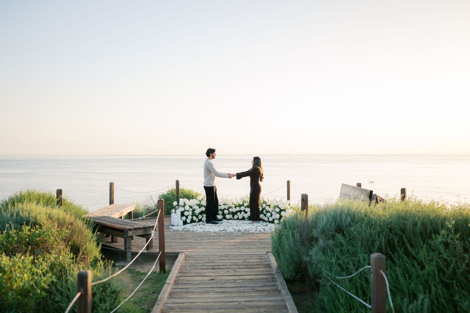 A series of wooden boardwalks lead through coastal dunes and beach grass to the ocean at sunset.