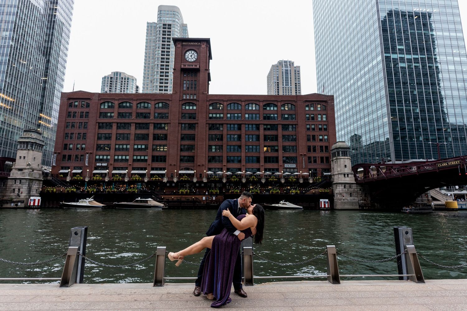 People enjoy views of Boston Harbor and city skyline from a waterfront promenade on a sunny day.