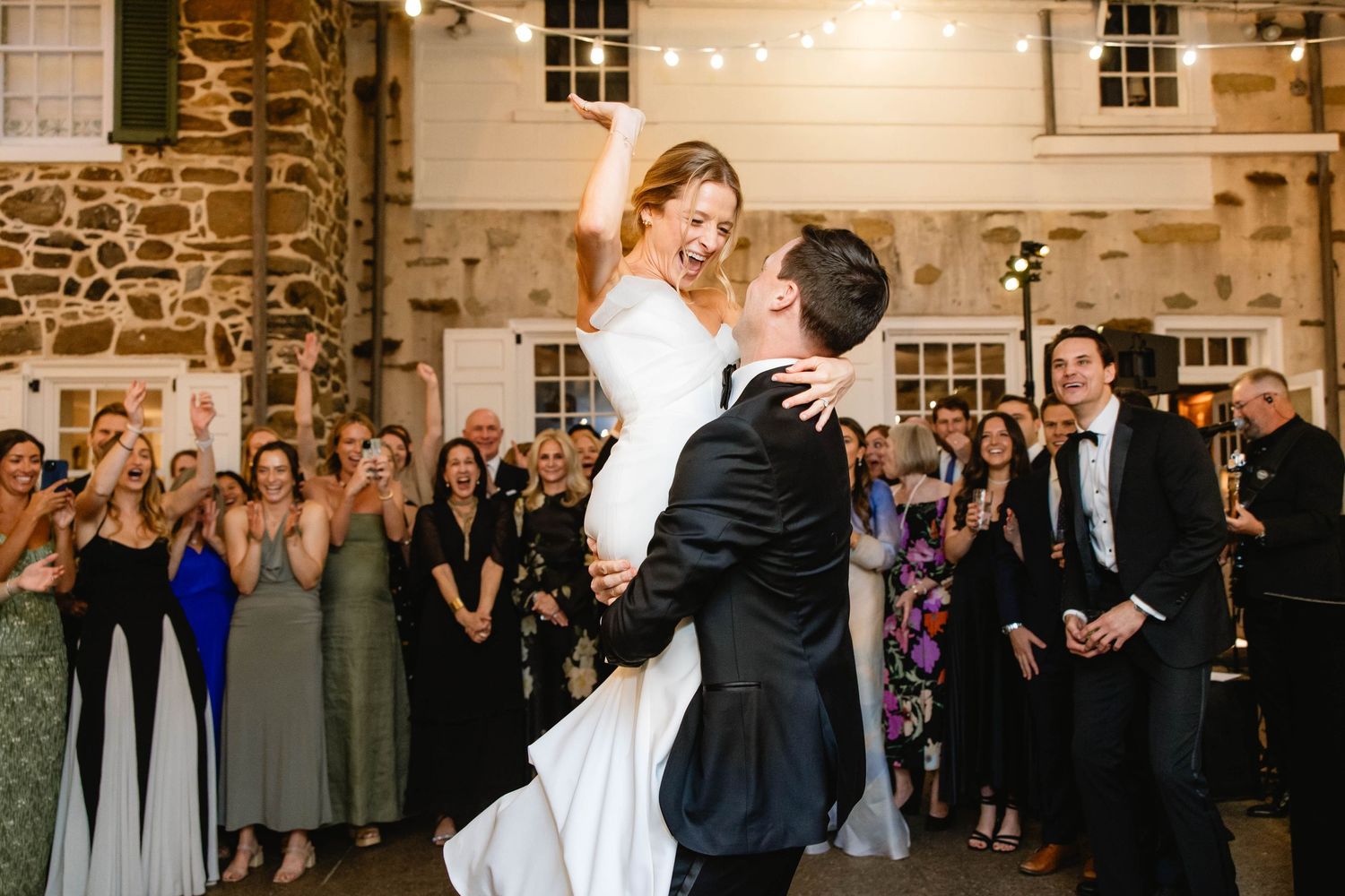 Wedding guests gather in rustic venue with exposed stone walls and string lights for first dance. Appleford Estate. Villanova, PA.