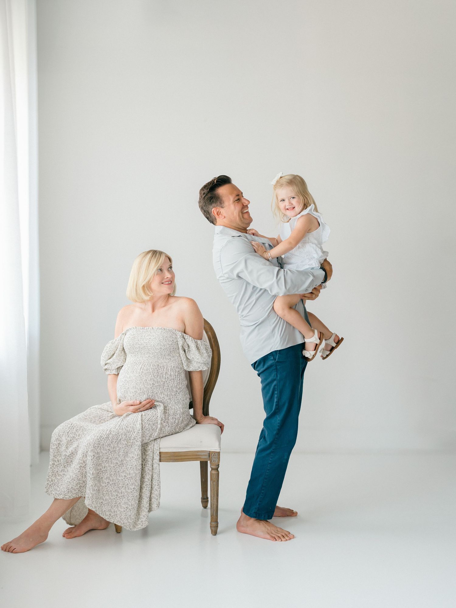 A father holds his daughter while her mother sits on a chair behind them smiling during their maternity photo session in Monmouth County NJ.