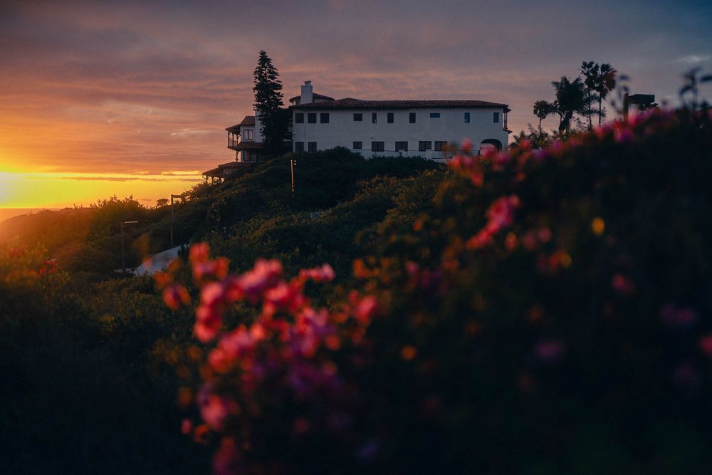 White Spanish-style house on hilltop at sunset with pink bougainvillea flowers in foreground silhouetted against orange sky.