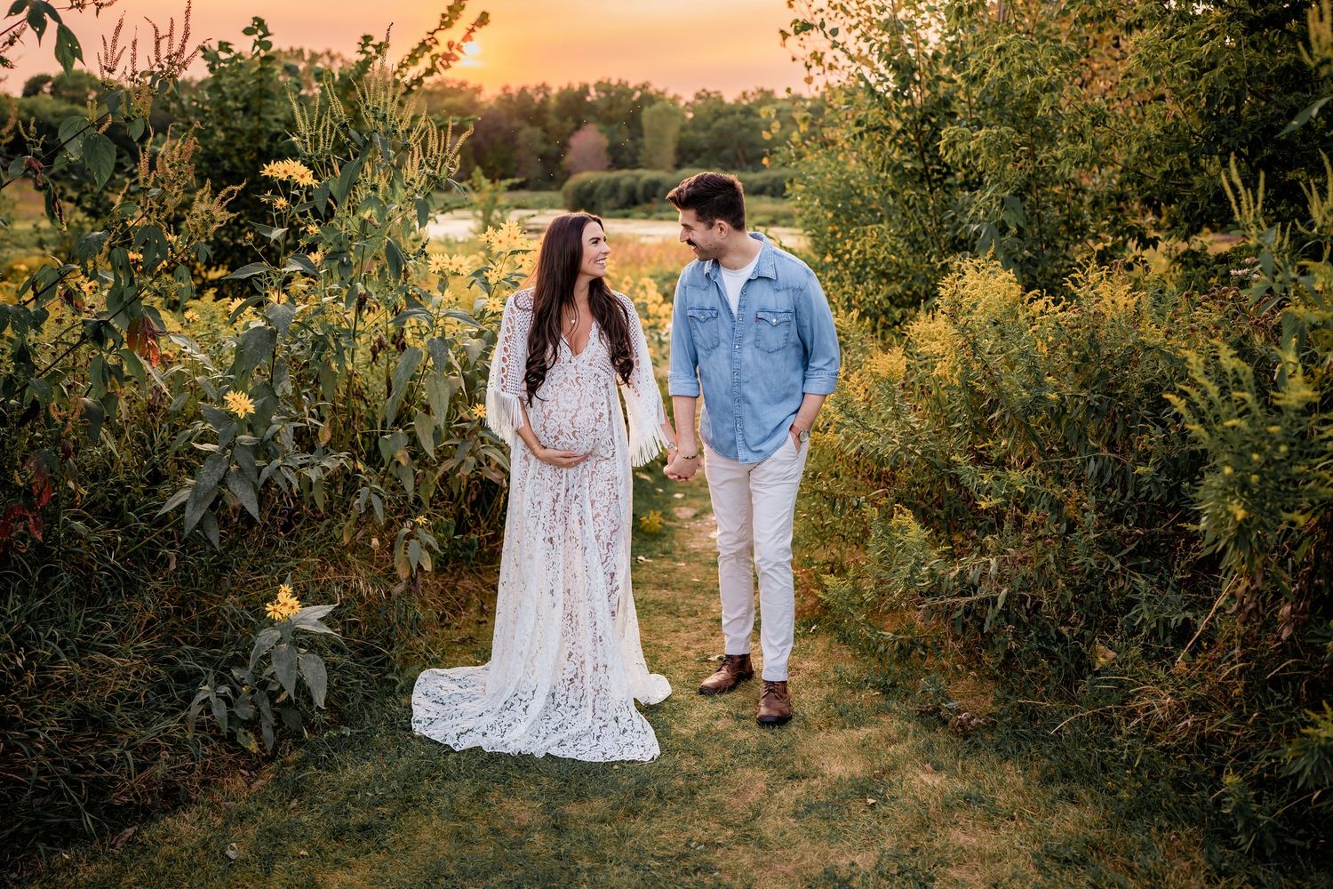 A woman in a burgundy dress and small girl in pink tulle dress hold hands on a path at golden sunset.