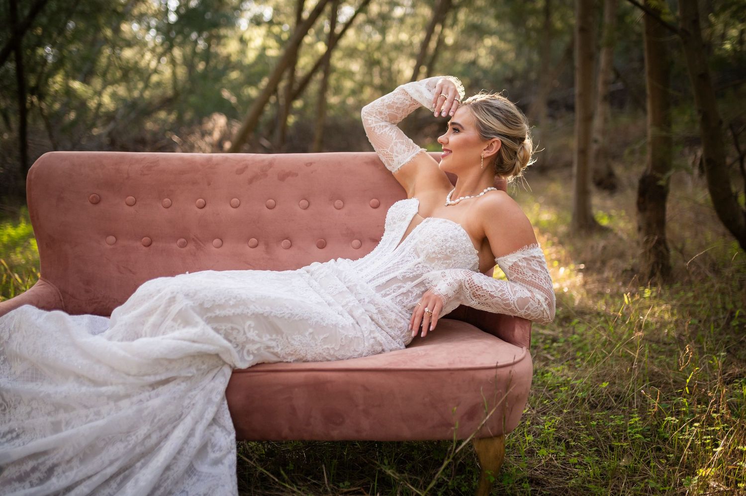 Elegant white lace wedding gown draped across dusty pink velvet sofa in sunlit forest setting.