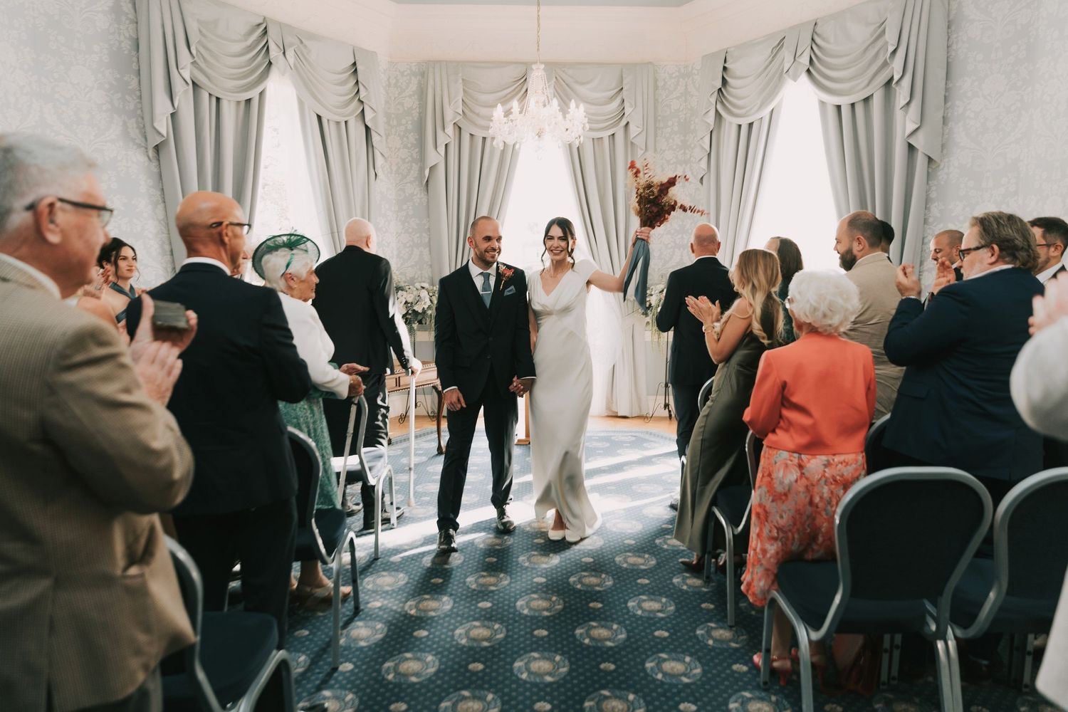 A wedding ceremony takes place in an elegant white room with guests seated in rows of chairs and ornate curtains.