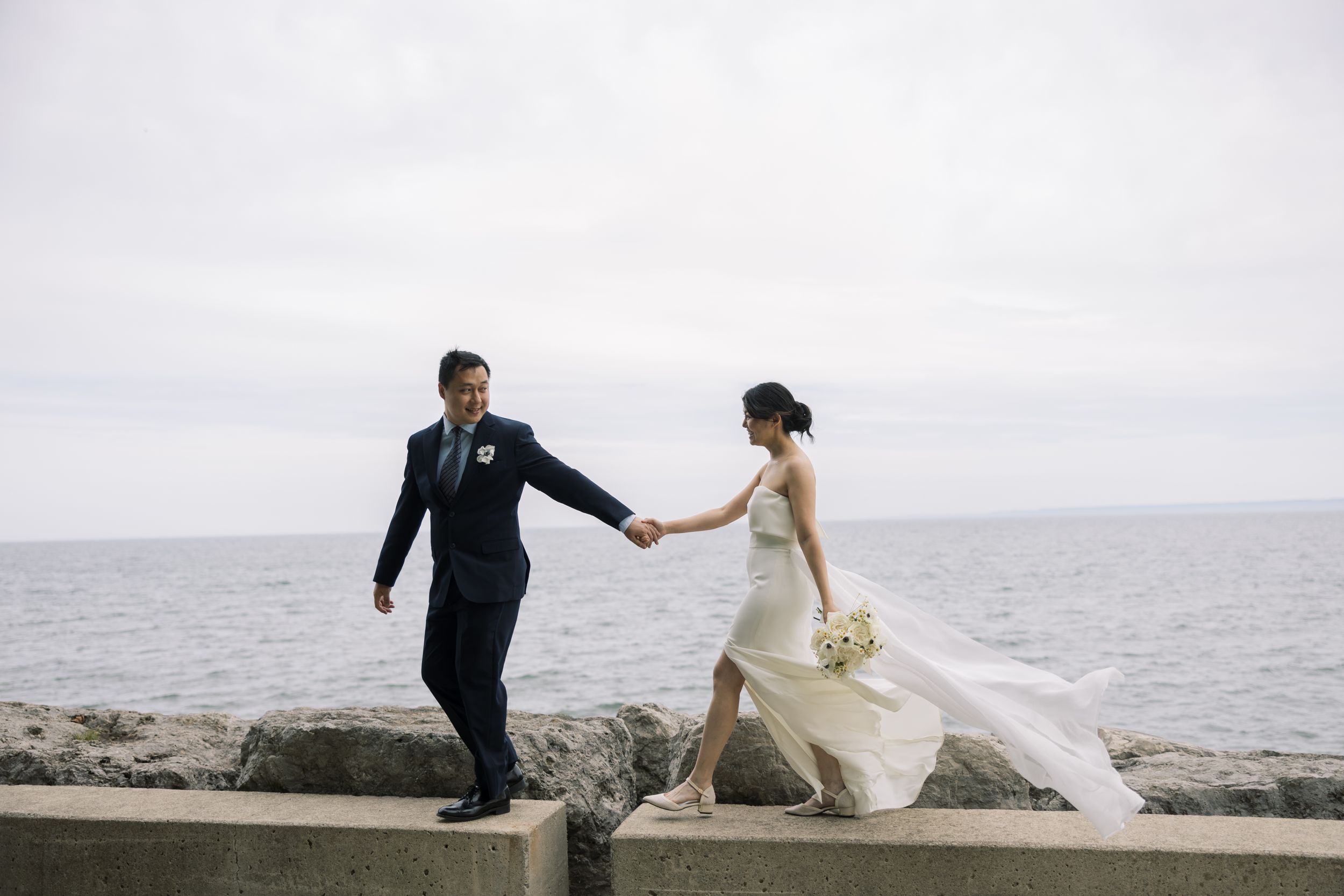 Newlyweds hold hands while walking along a rocky seawall with ocean waves in the background.