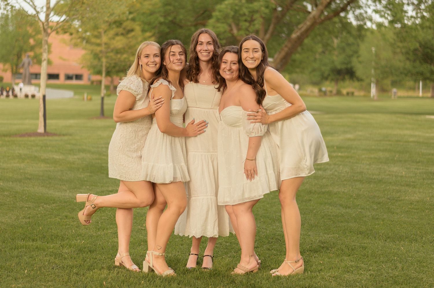 A group of friends in white dresses pose together on a sunny green lawn.