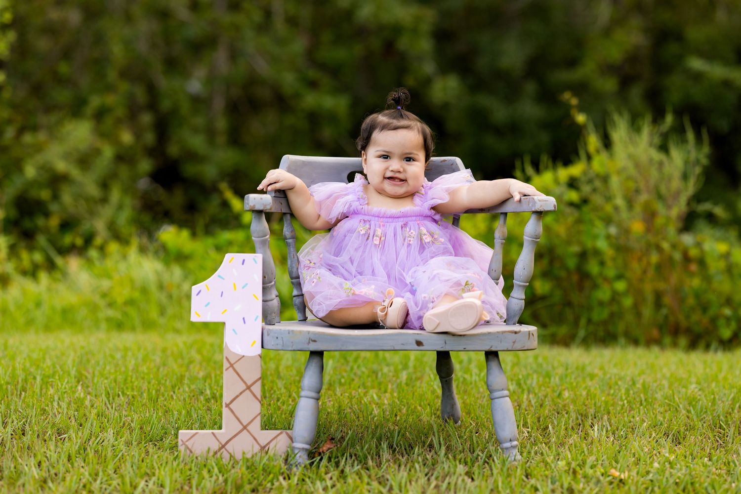 A baby in a pink dress and white tights lays on a wooden bench in a grassy outdoor setting.