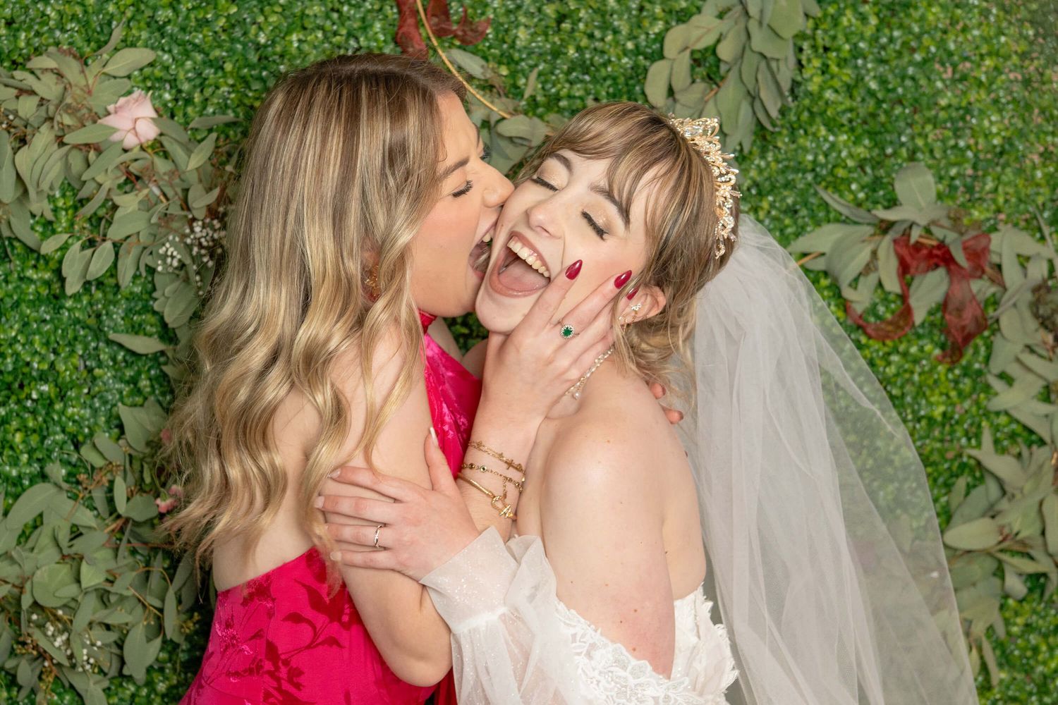 Two women share a joyful candid moment against a green garden backdrop, one wearing a wedding dress with veil.