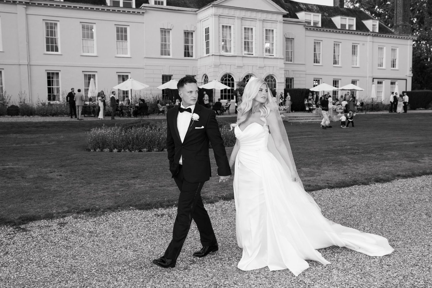 A wedding couple walks along a gravel path outside an elegant white mansion estate on their special day.