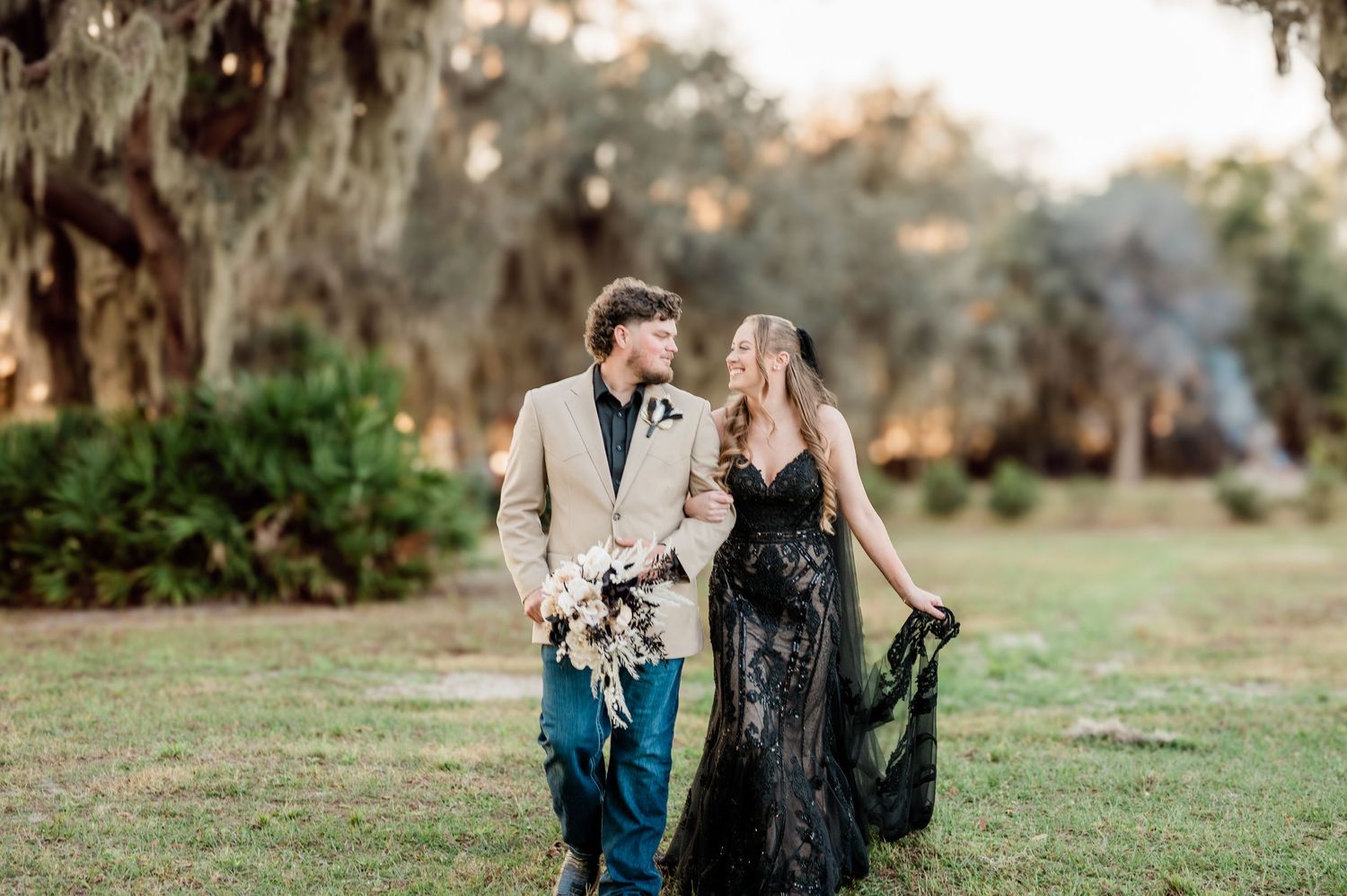 A couple in formal attire shares a romantic moment on a grassy lawn beneath Spanish moss-draped trees at sunset.