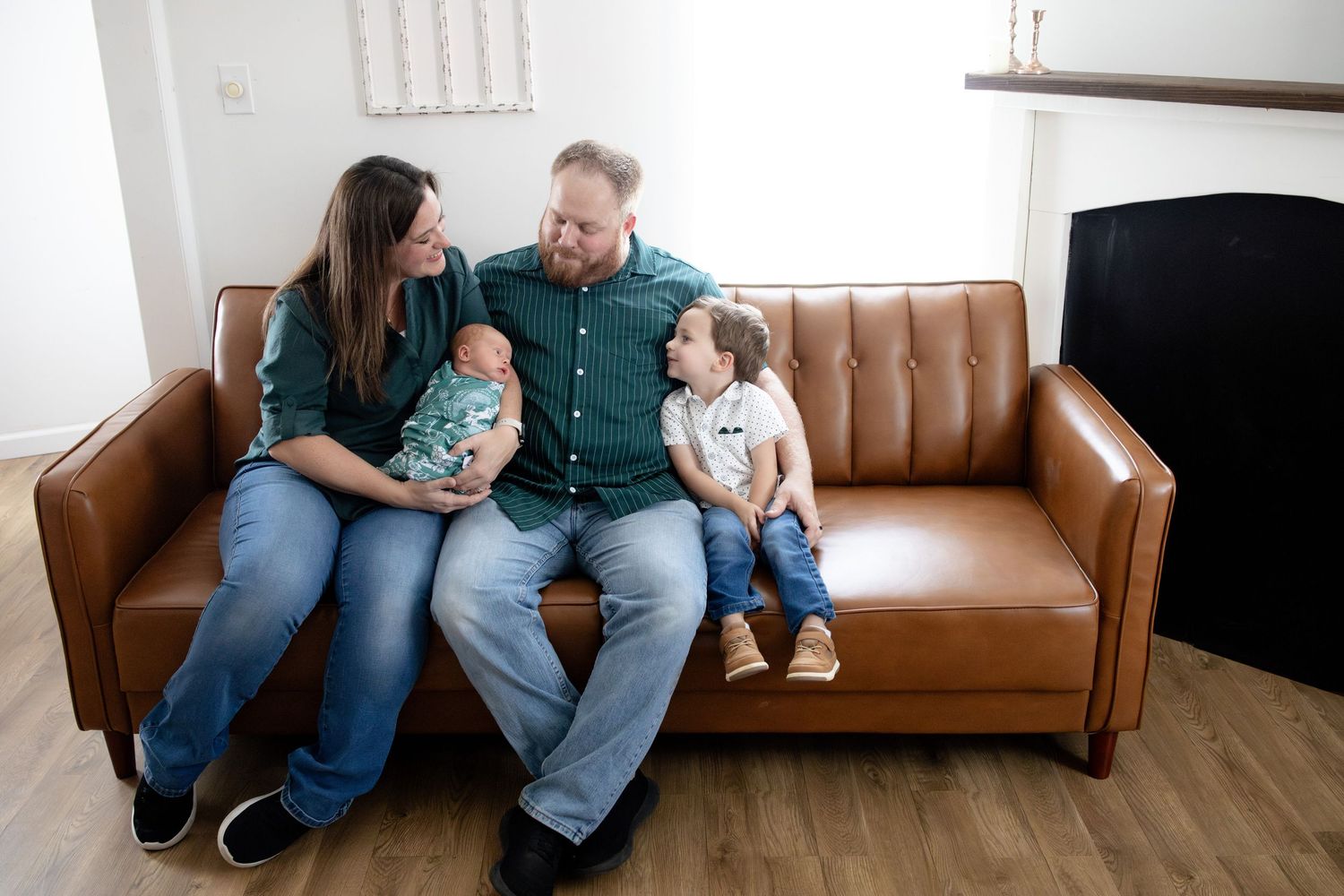 Family of four sits on brown leather sofa, parents smiling at newborn baby while toddler sits beside them.
