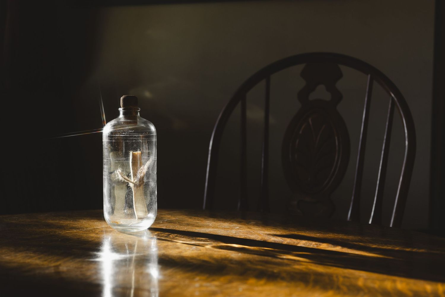 A clear glass bottle with a cork stopper sits on a wooden table beside a dark wooden chair in moody lighting.