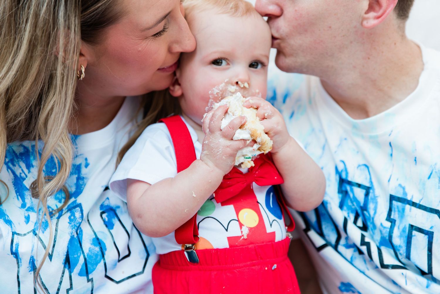 Parents kissing their baby on cheeks while toddler eats birthday cake smash in red overalls.