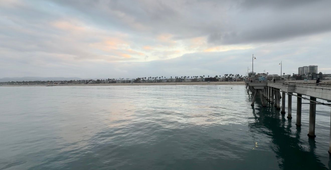 Calm ocean waters beside a wooden pier at dusk with a cloudy pastel sky and distant shoreline with palm trees.
