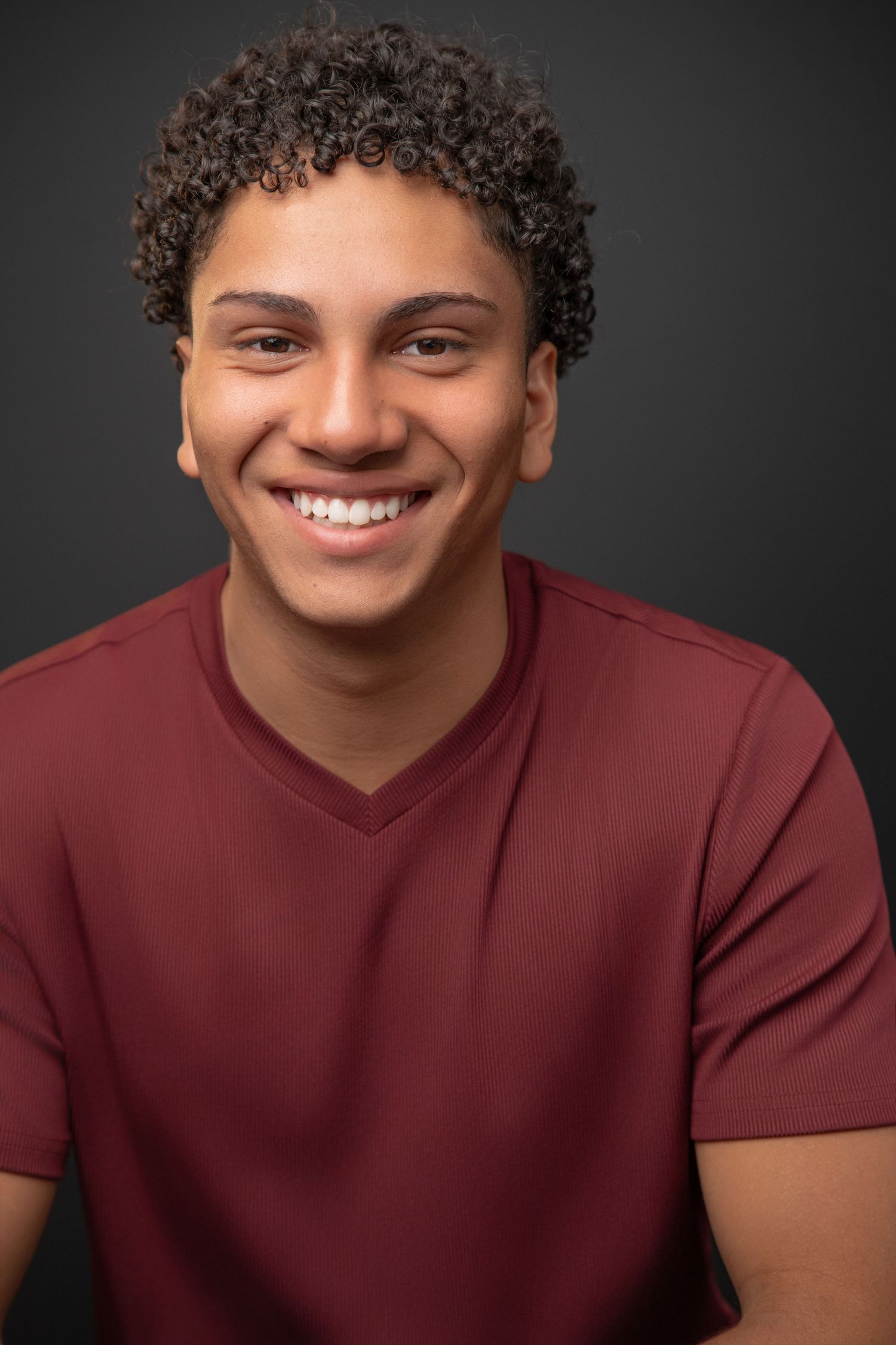 Young man with curly hair smiling confidently in a teal henley shirt against a gray background.