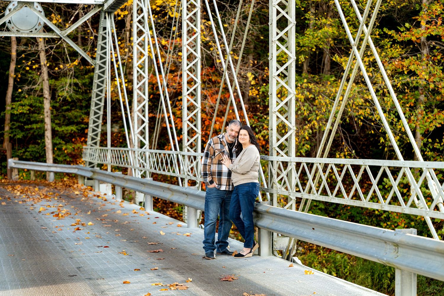 Couple embracing on a historic steel truss bridge surrounded by colorful autumn foliage and fallen leaves.