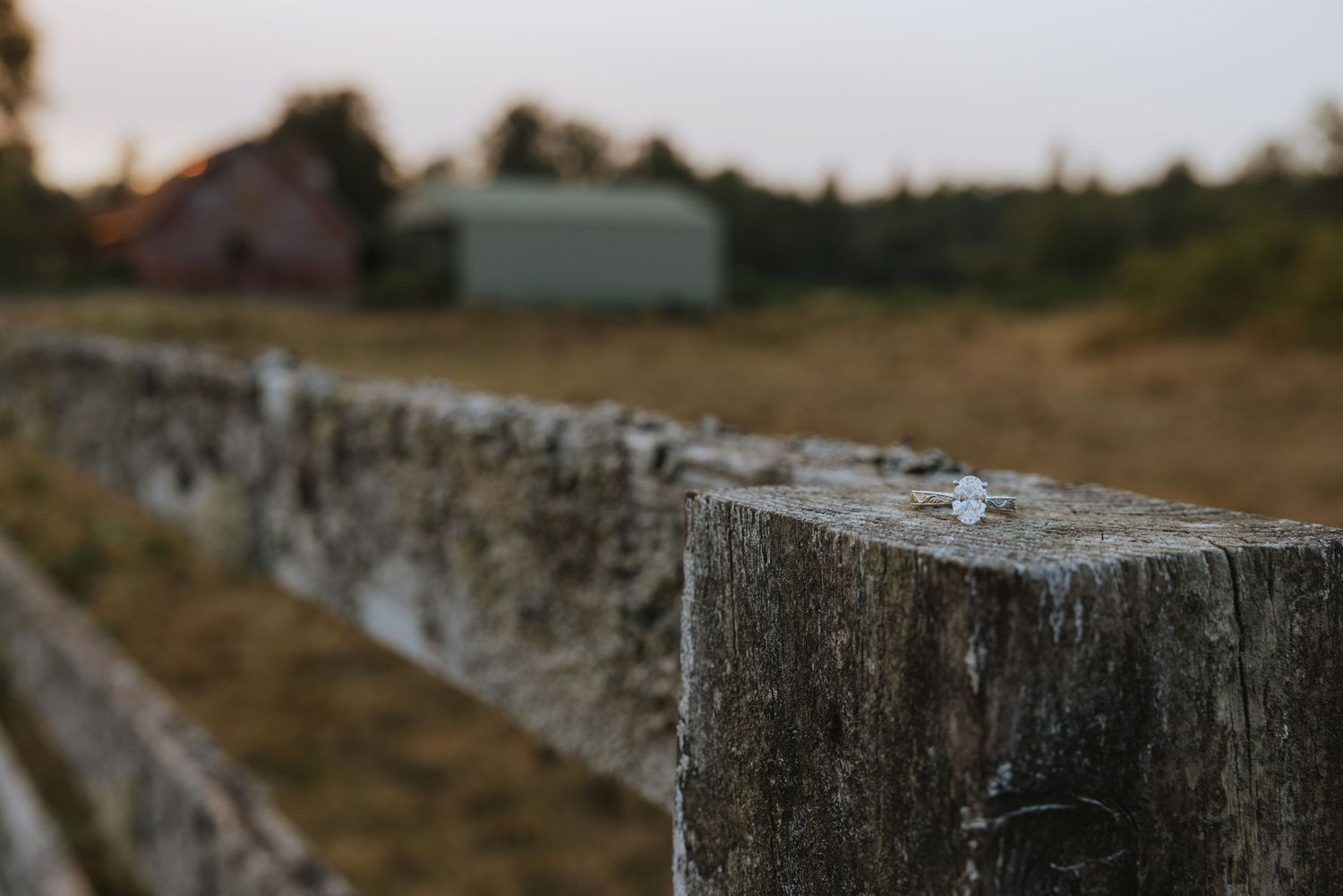Rustic wooden fence post in foreground with blurred farm buildings and trees at dusk in rural countryside.