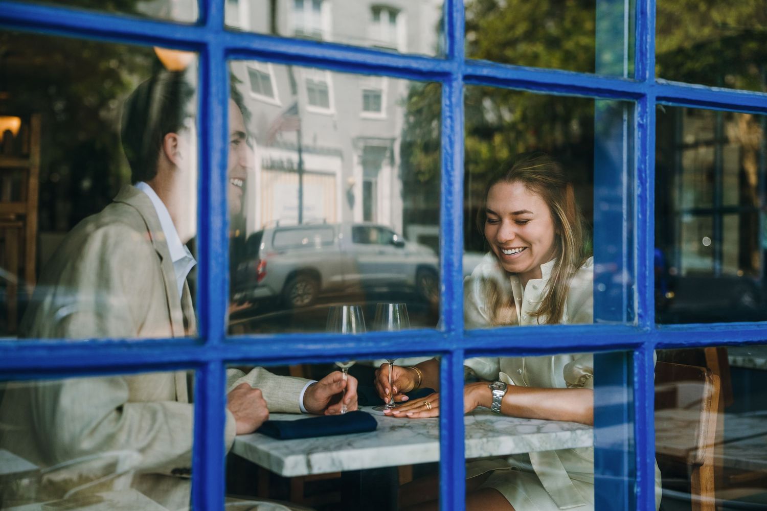 Couple smiling and sharing a meal together at a cafe table, viewed through a blue-framed window.