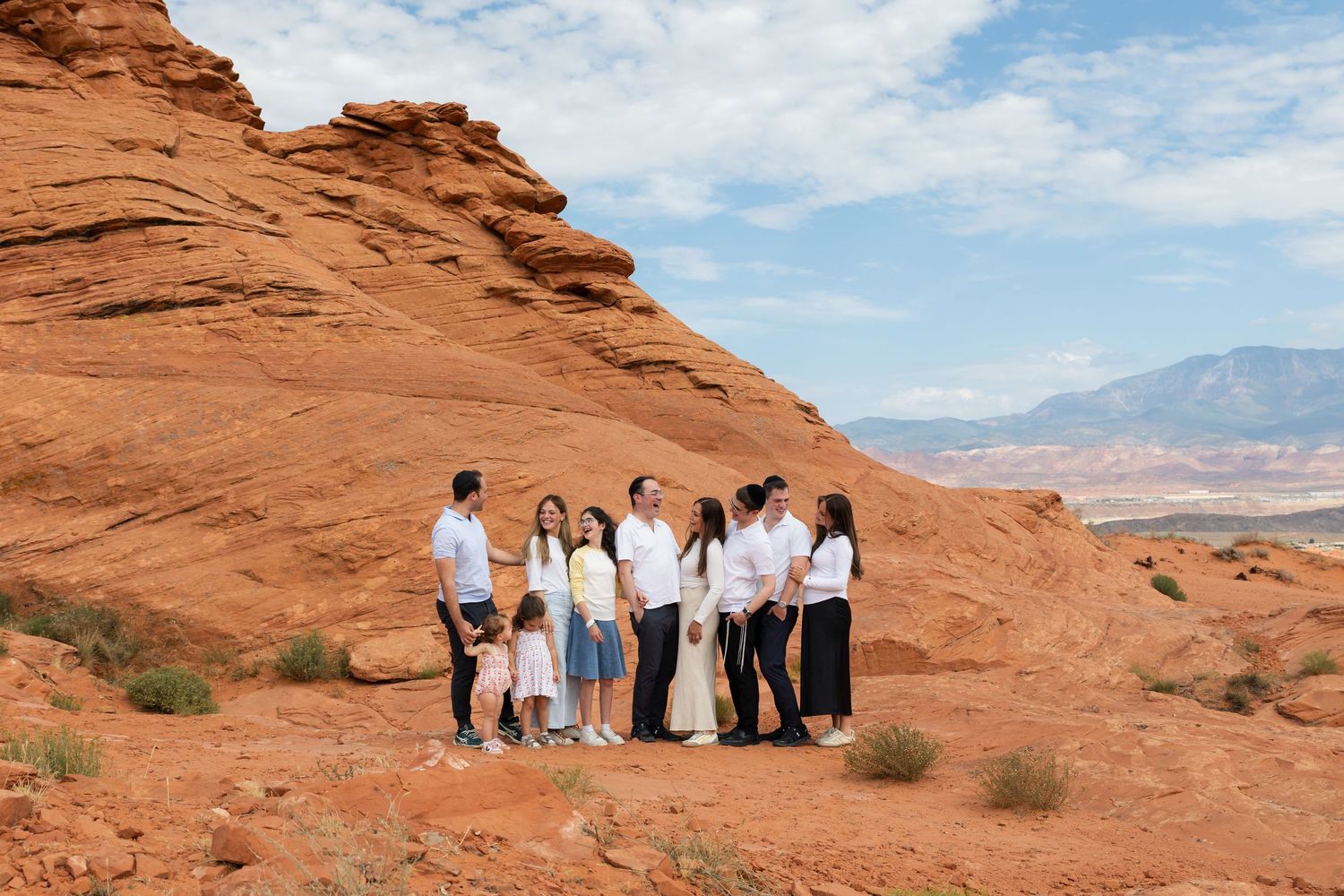 Family group photo session among red rock formations in a desert landscape with mountains and blue sky backdrop.